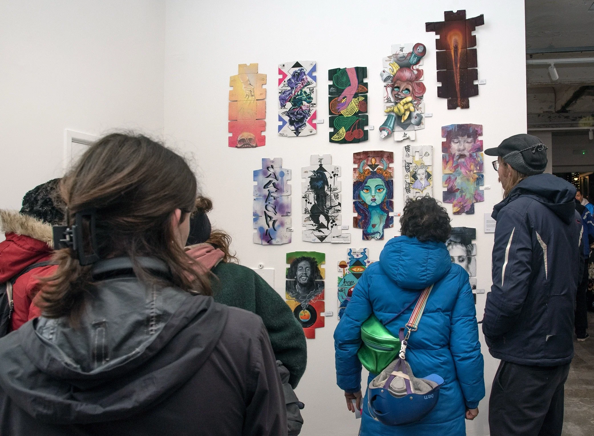 Visitors at a gallery view a white wall hung with artworks painted on flattened Loop spray can boxes from "The Queens of the Wall" at Copeland Park, Peckham in 2024. The cardboard boxes feature graffiti lettering, flowers in street art styles.