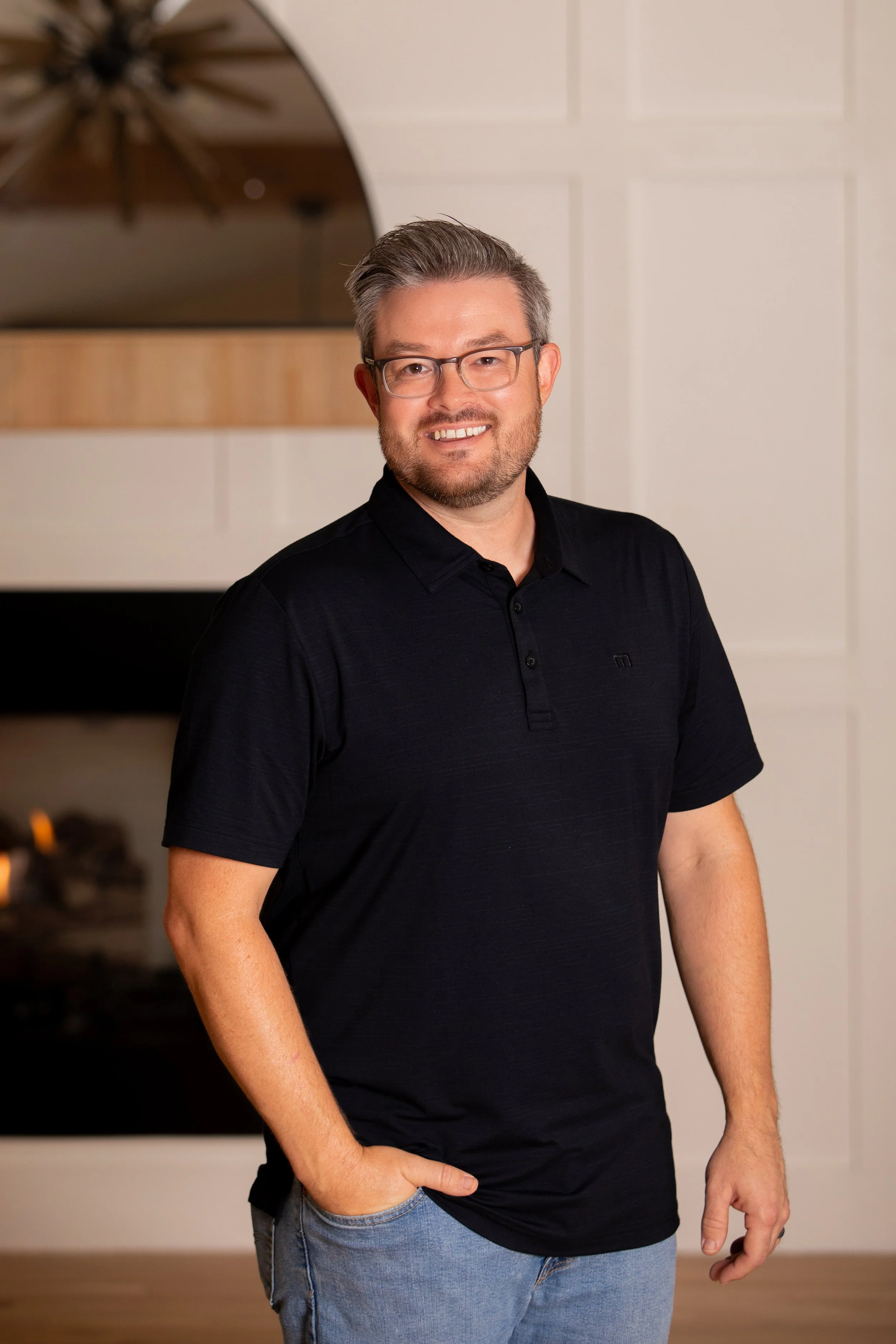 A man with gray hair, glasses, and a beard stands indoors, smiling. He is wearing a black short-sleeved polo shirt and light blue jeans, with his right hand in his pocket.