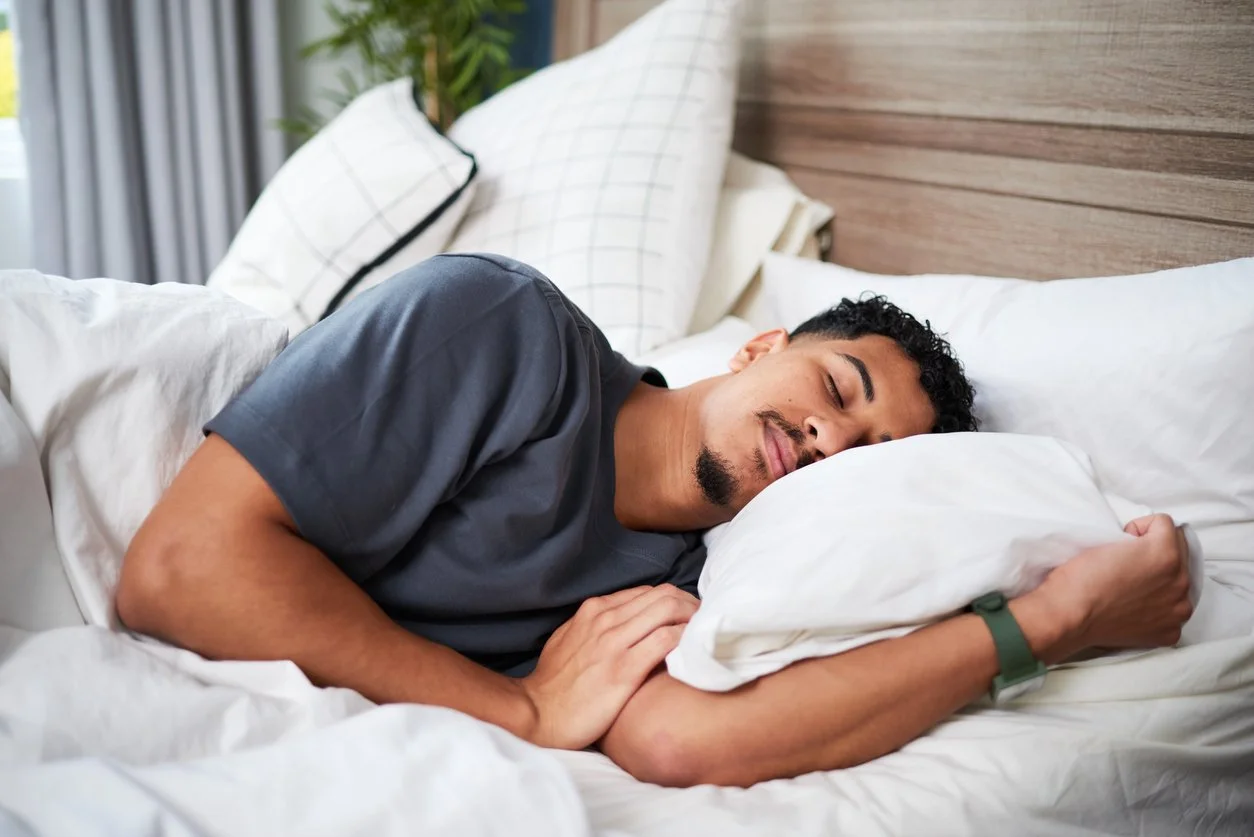 A man with short curly hair and a goatee sleeping on his side with a pillow in a bedroom with a wooden headboard and checkered pillows.