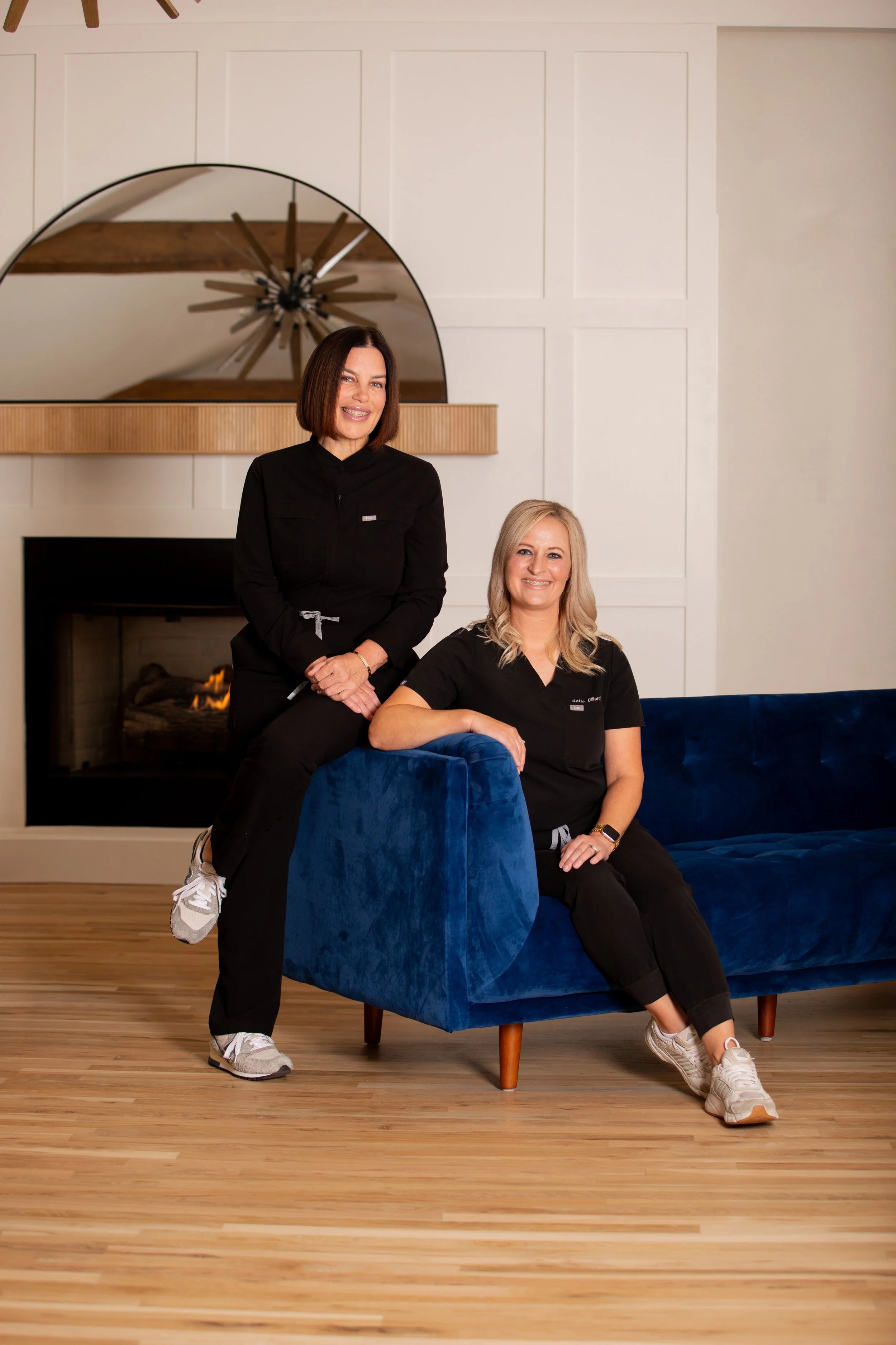 Two women in black scrubs sitting and standing on a blue couch in front of a fireplace, with a mirror and modern decor behind them.