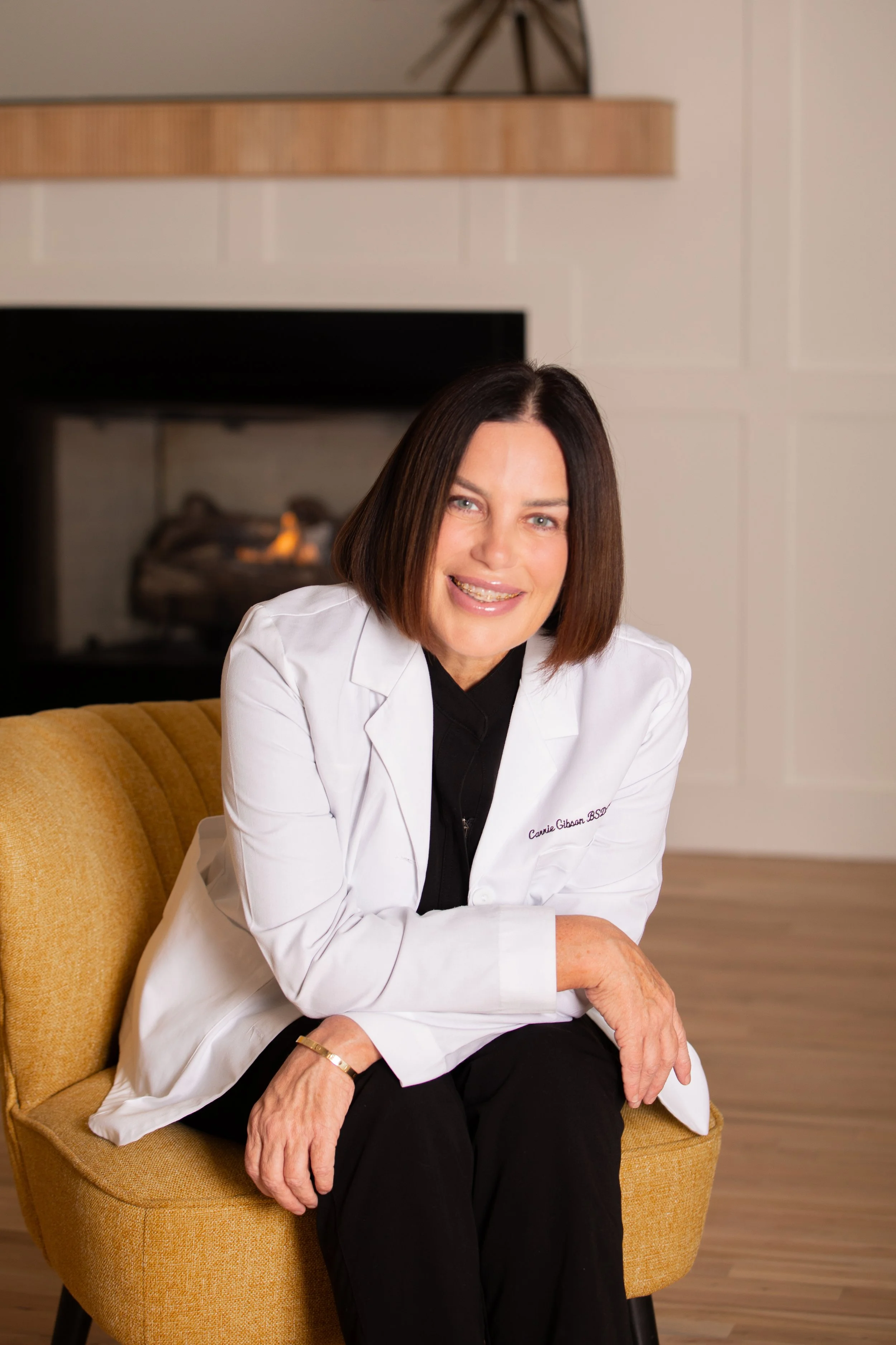A woman with shoulder-length brown hair smiling, wearing a white doctor's coat with embroidered name, sitting on a yellow chair in a cozy living room with a fireplace.