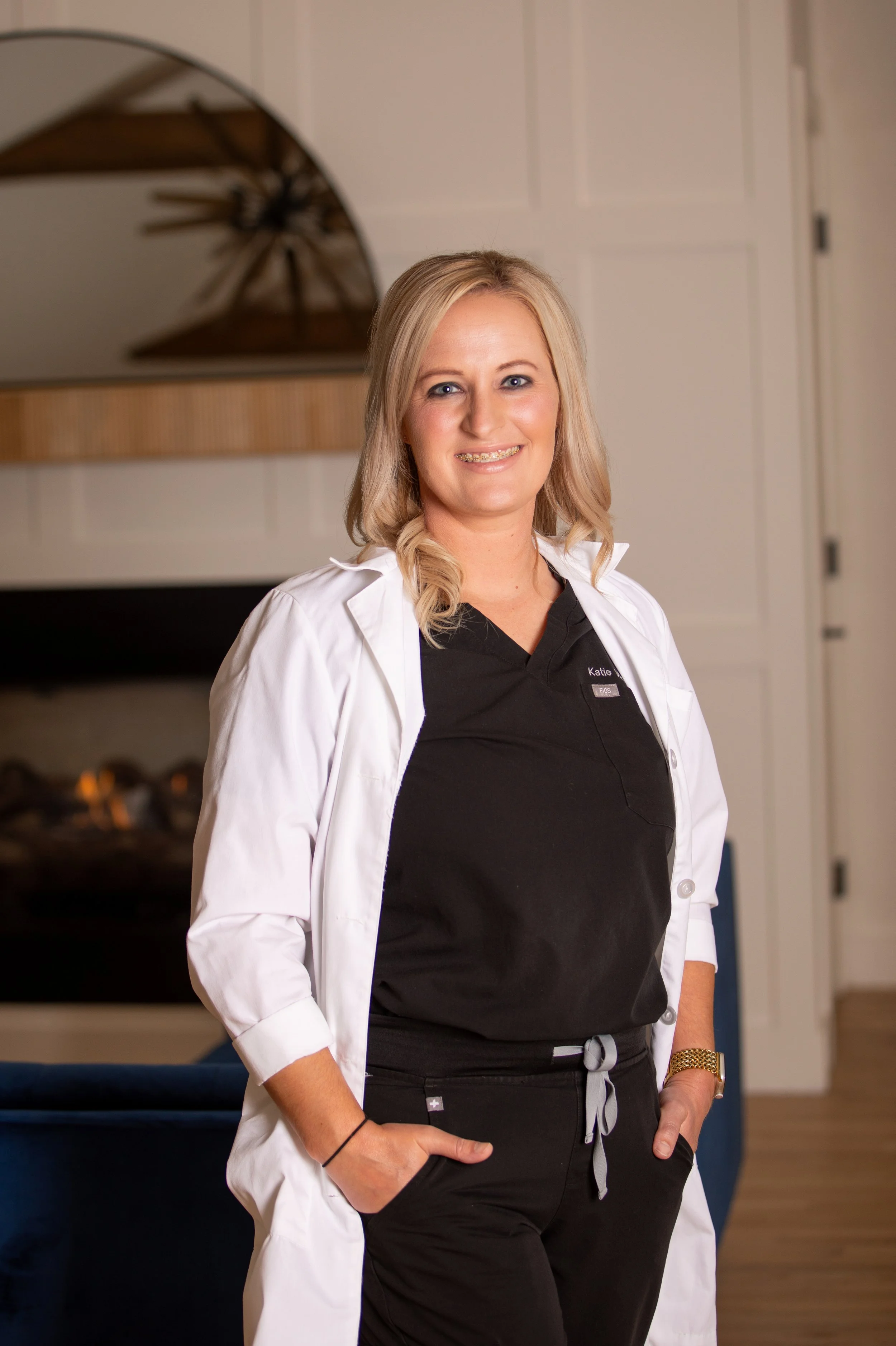 A woman in medical scrubs and a white lab coat standing indoors with her hands in her pockets, smiling at the camera.