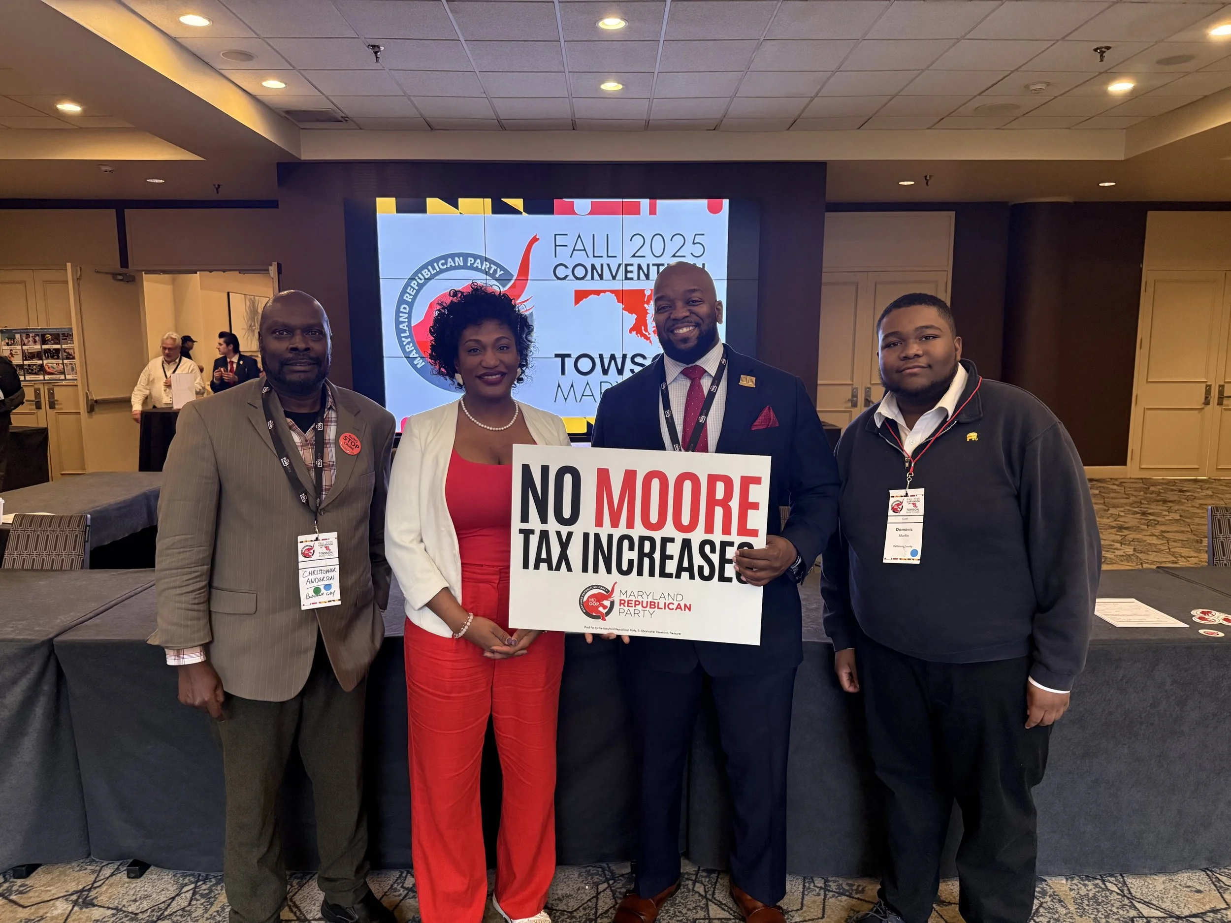 Four people standing together at a political event, holding a sign that says 'No Moore Tax Increases' with the Maryland Republican Party logo. They are in a conference room with a large screen behind them displaying the Maryland Republican Party logo and the words 'Fall 2025 Convention.'