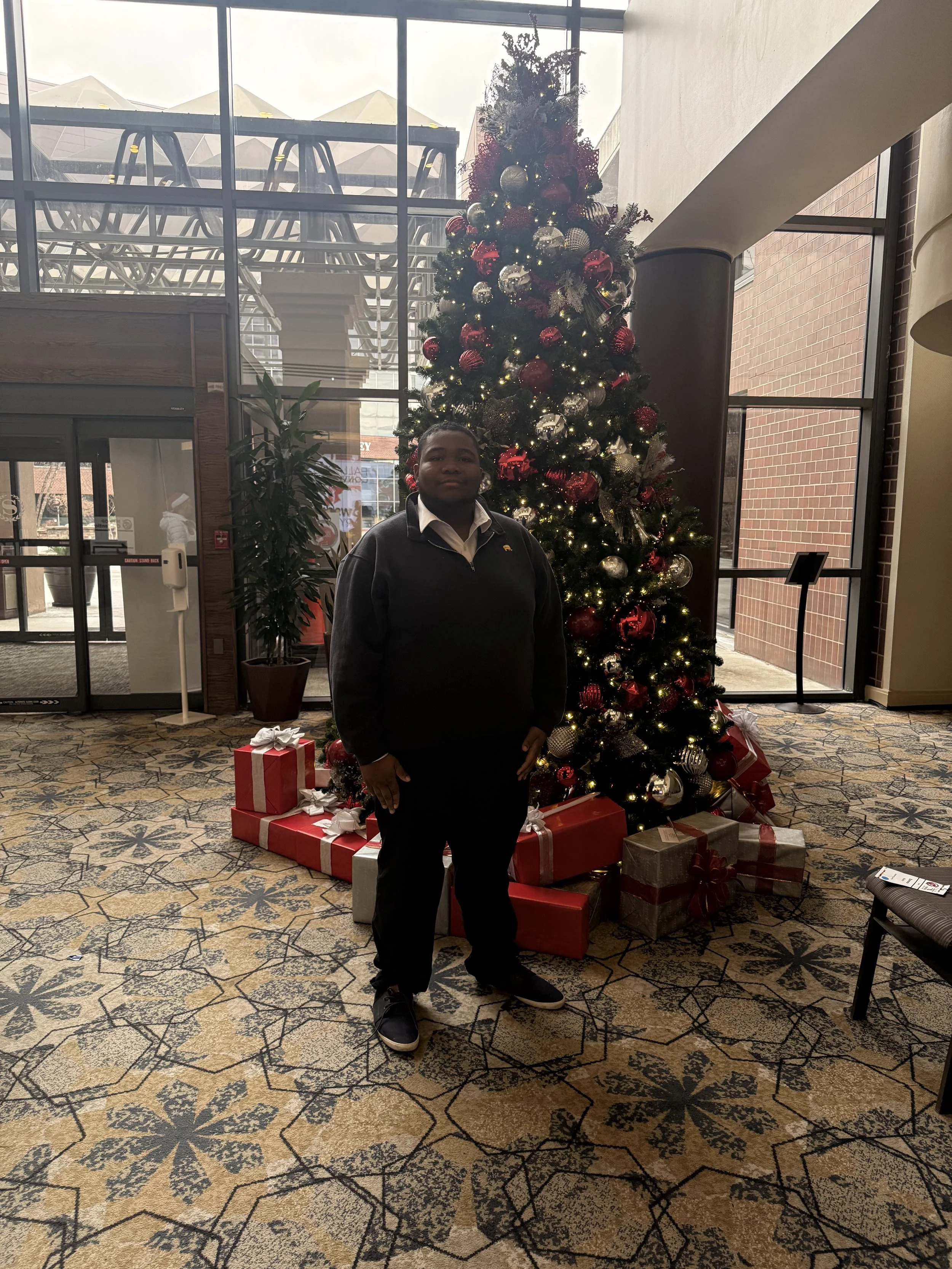 A person standing in front of a decorated Christmas tree with presents underneath, inside a building with large windows.