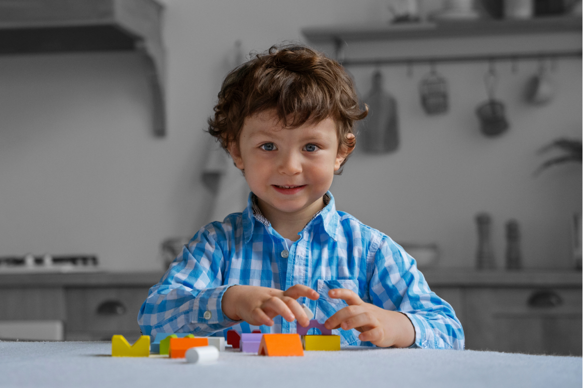 A young boy with curly brown hair and blue eyes, wearing a blue checkered shirt, sitting at a table with colorful alphabet blocks.
