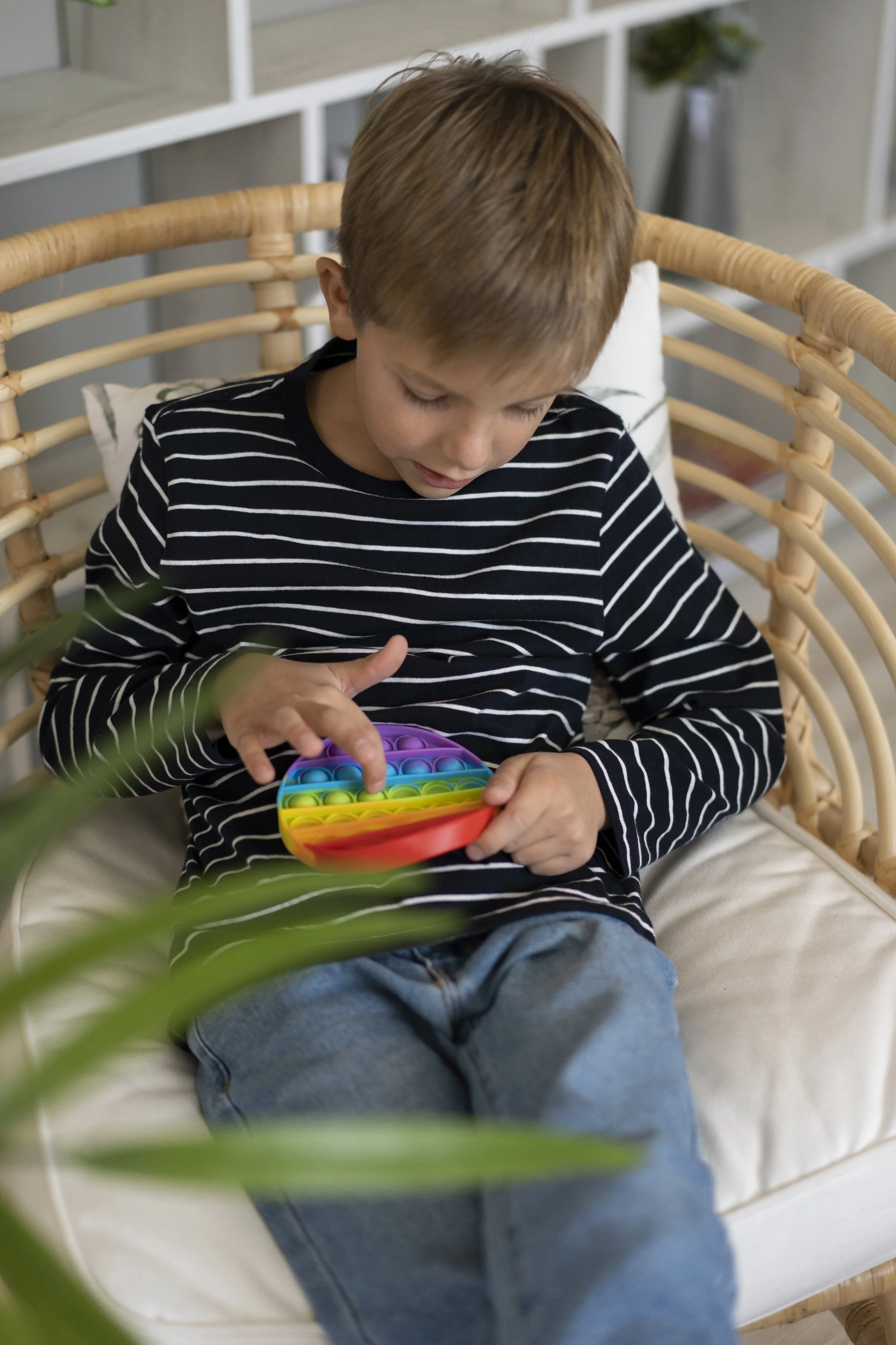 A young boy with light brown hair wearing a black and white striped shirt and jeans, sitting on a wicker chair, playing with a colorful rainbow-colored pop-it fidget toy.
