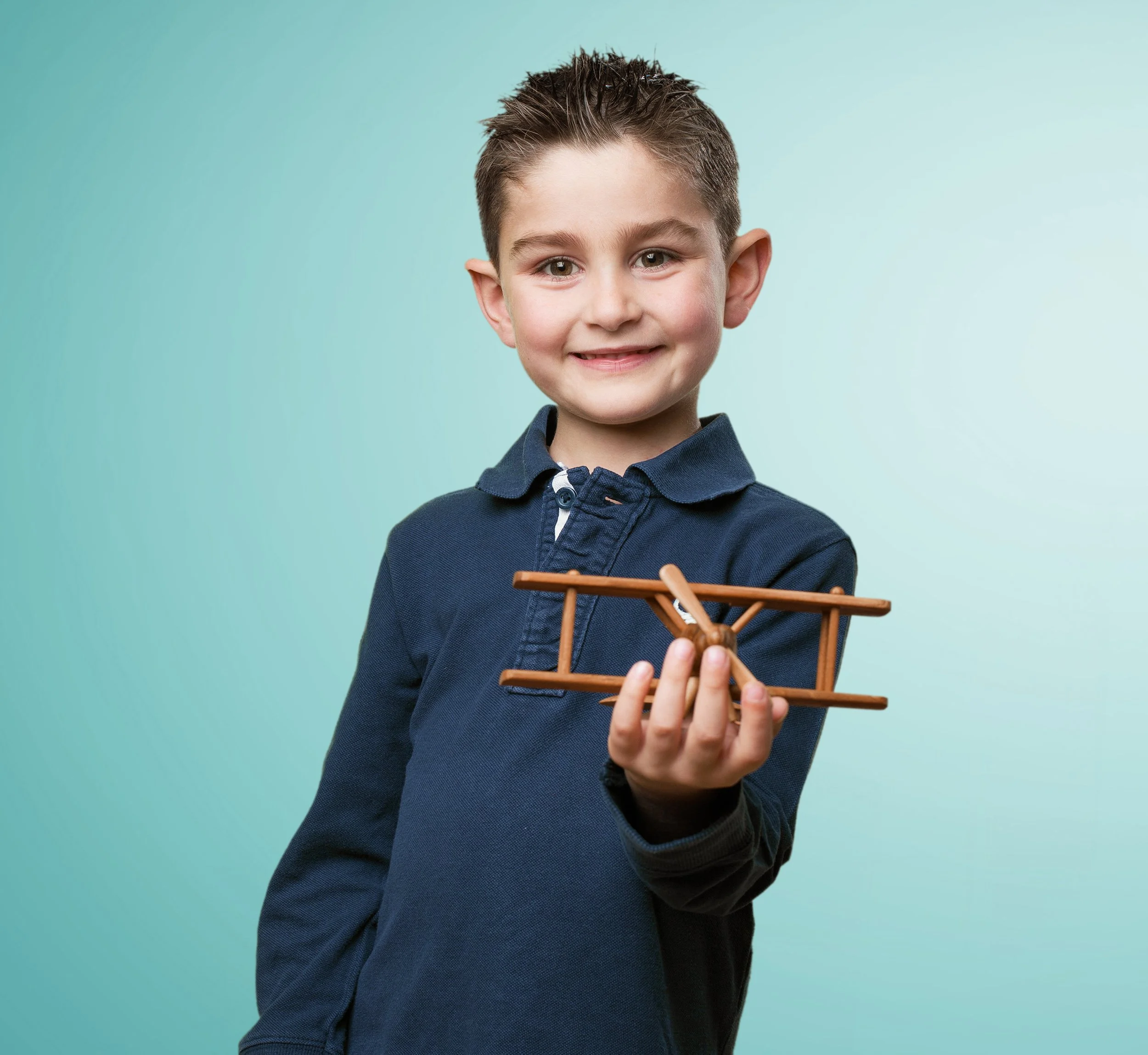 A young boy with short brown hair and a blue polo shirt smiling and holding a wooden model airplane against a light blue background.