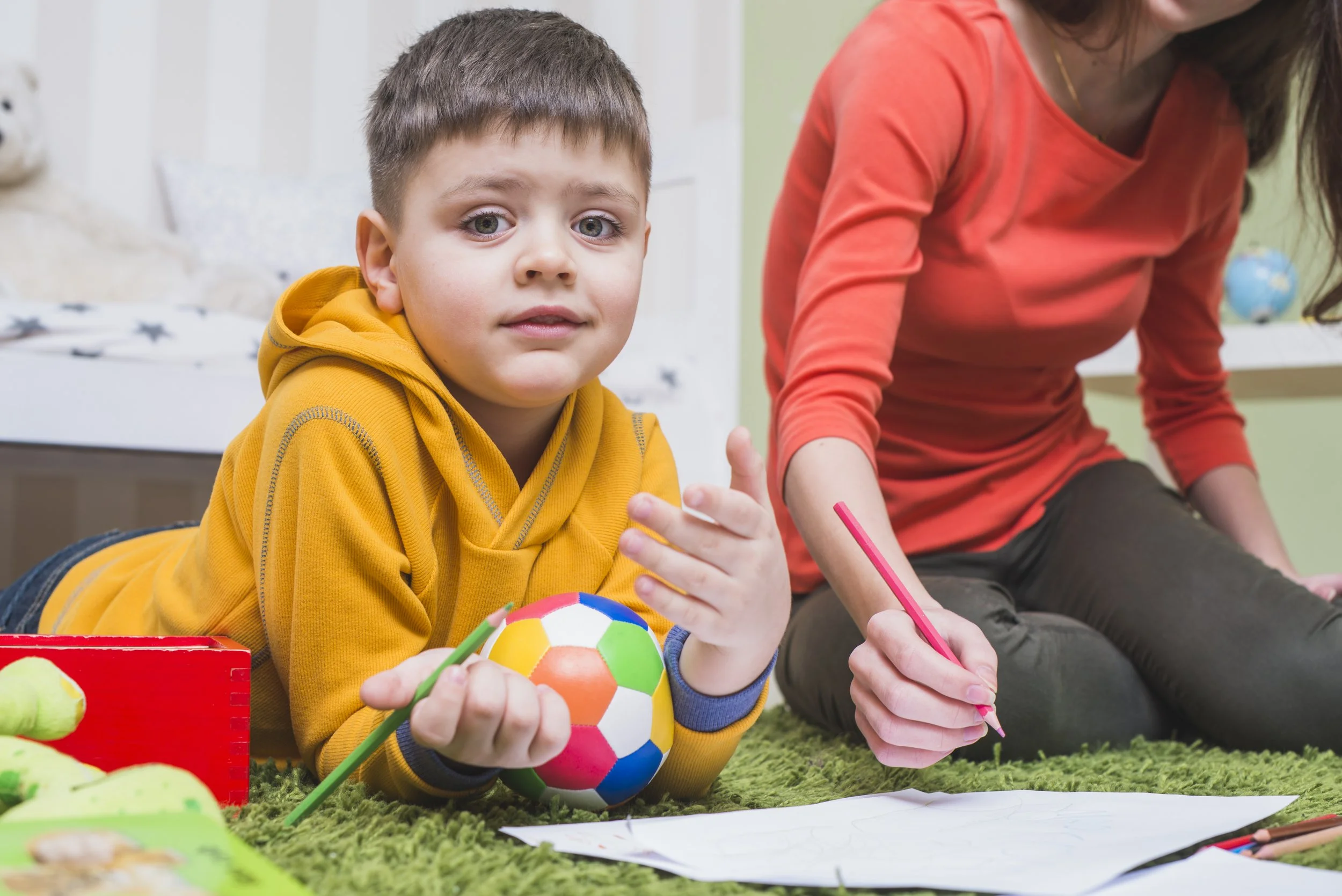 Young boy in a yellow hoodie lying on a green carpet, holding a colorful soccer ball, with papers and colored pencils nearby, and a woman in a red shirt sitting next to him.