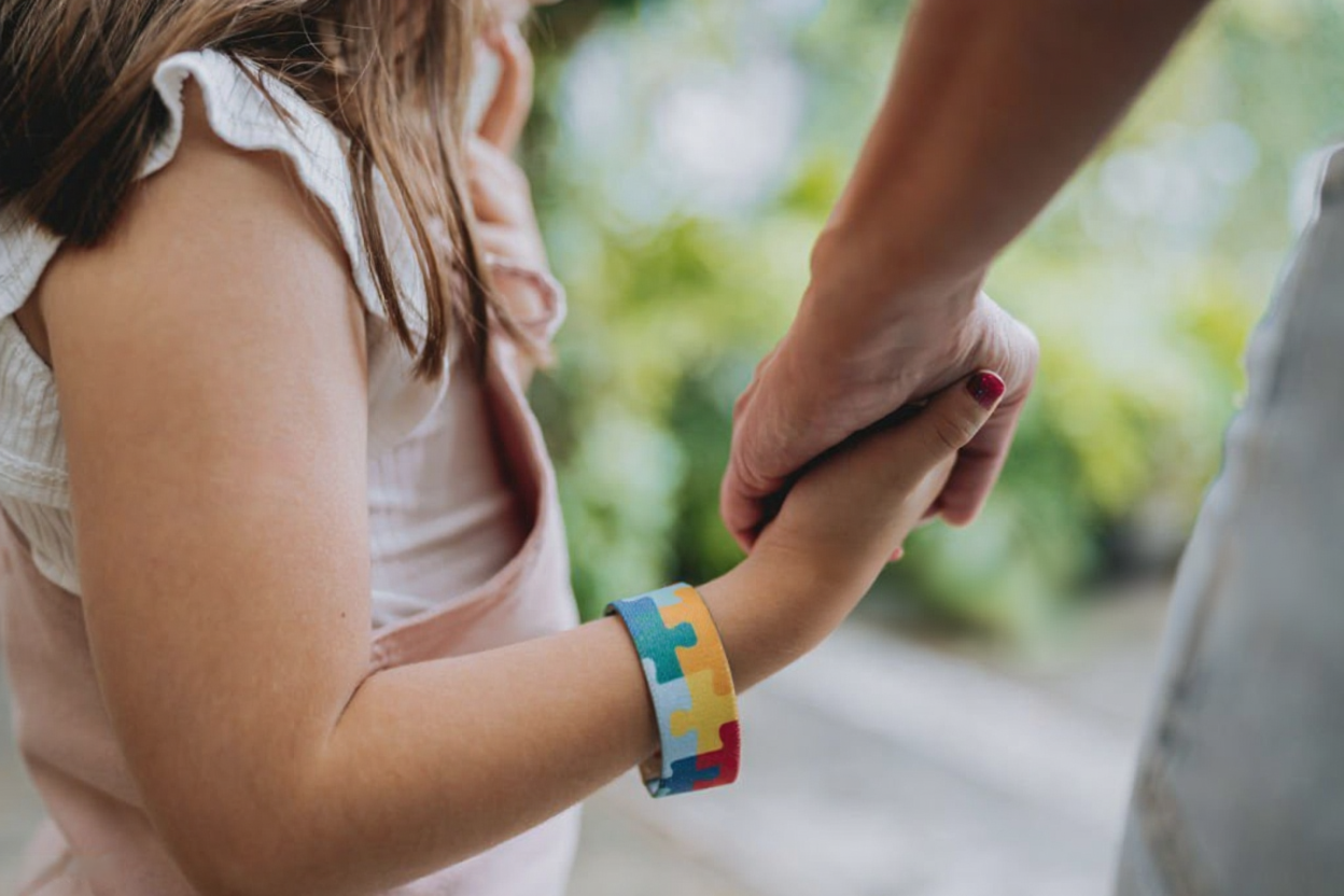 A young girl holding an adult's hand outdoors, wearing a colorful rubber bracelet with puzzle pieces, with a blurred green background.