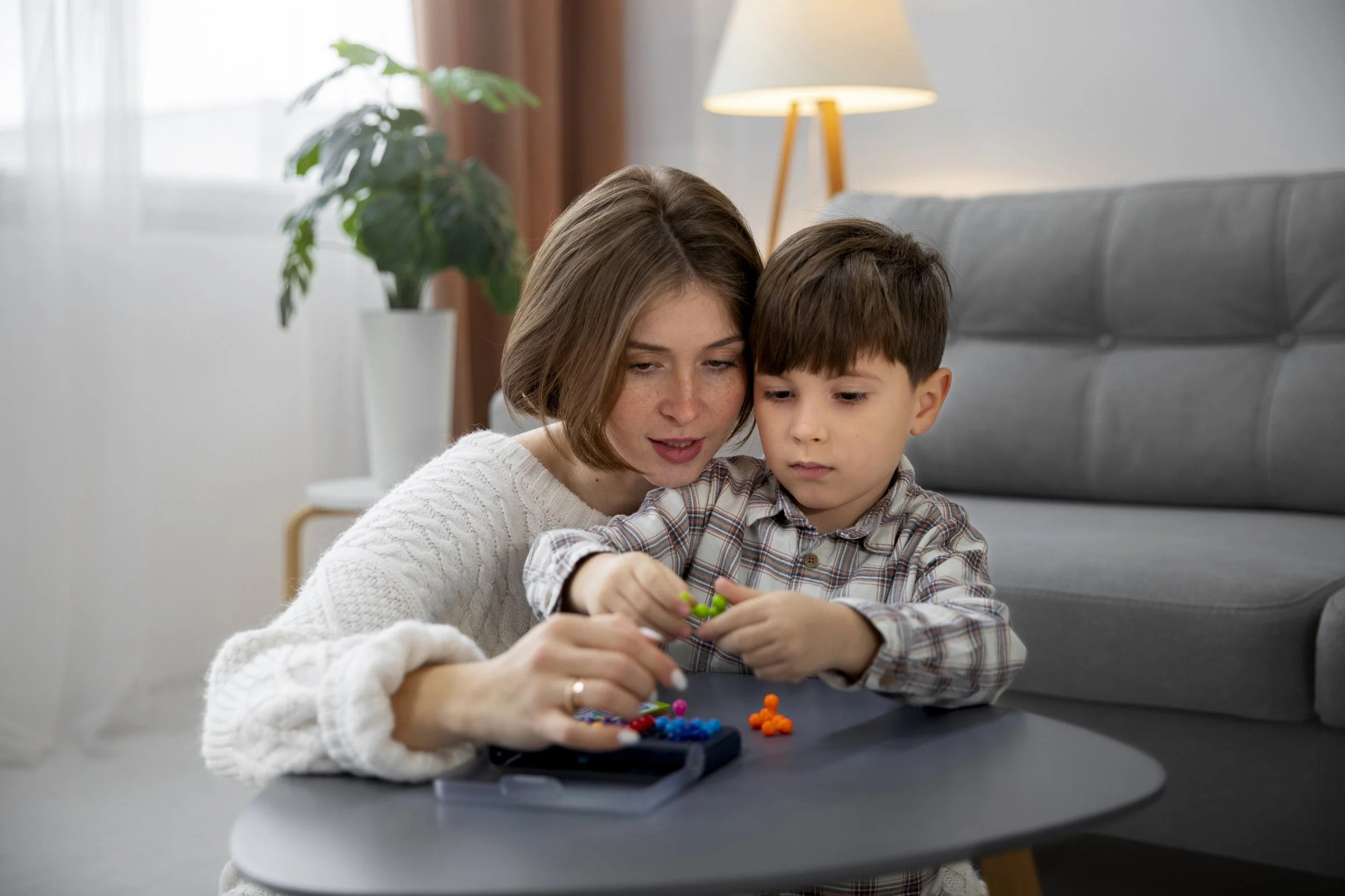 A woman and young boy playing a game of Connect 4 at a table in a cozy living room.