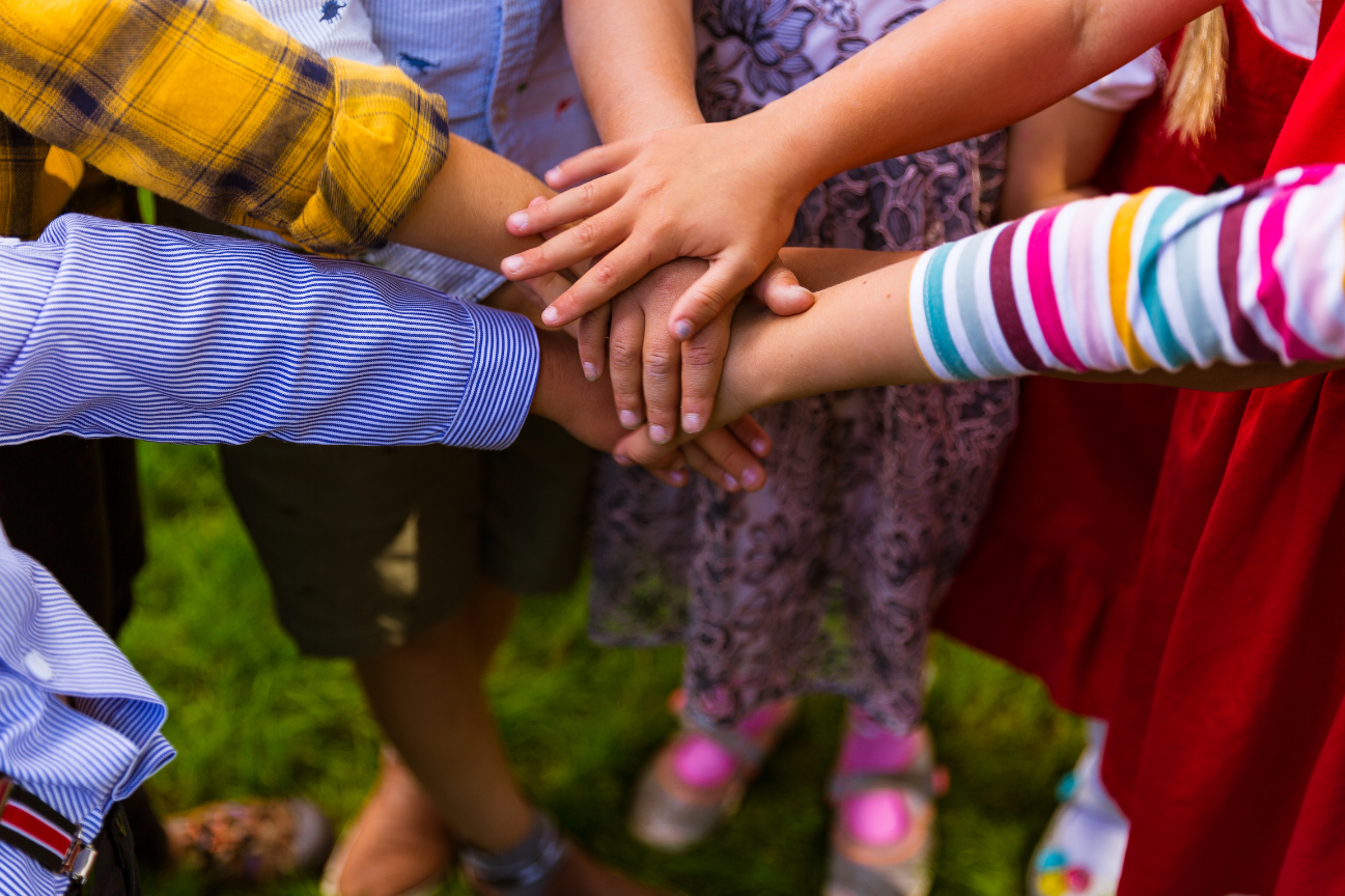 Children of diverse ethnicities join hands in a circle outdoors, symbolizing teamwork and friendship.