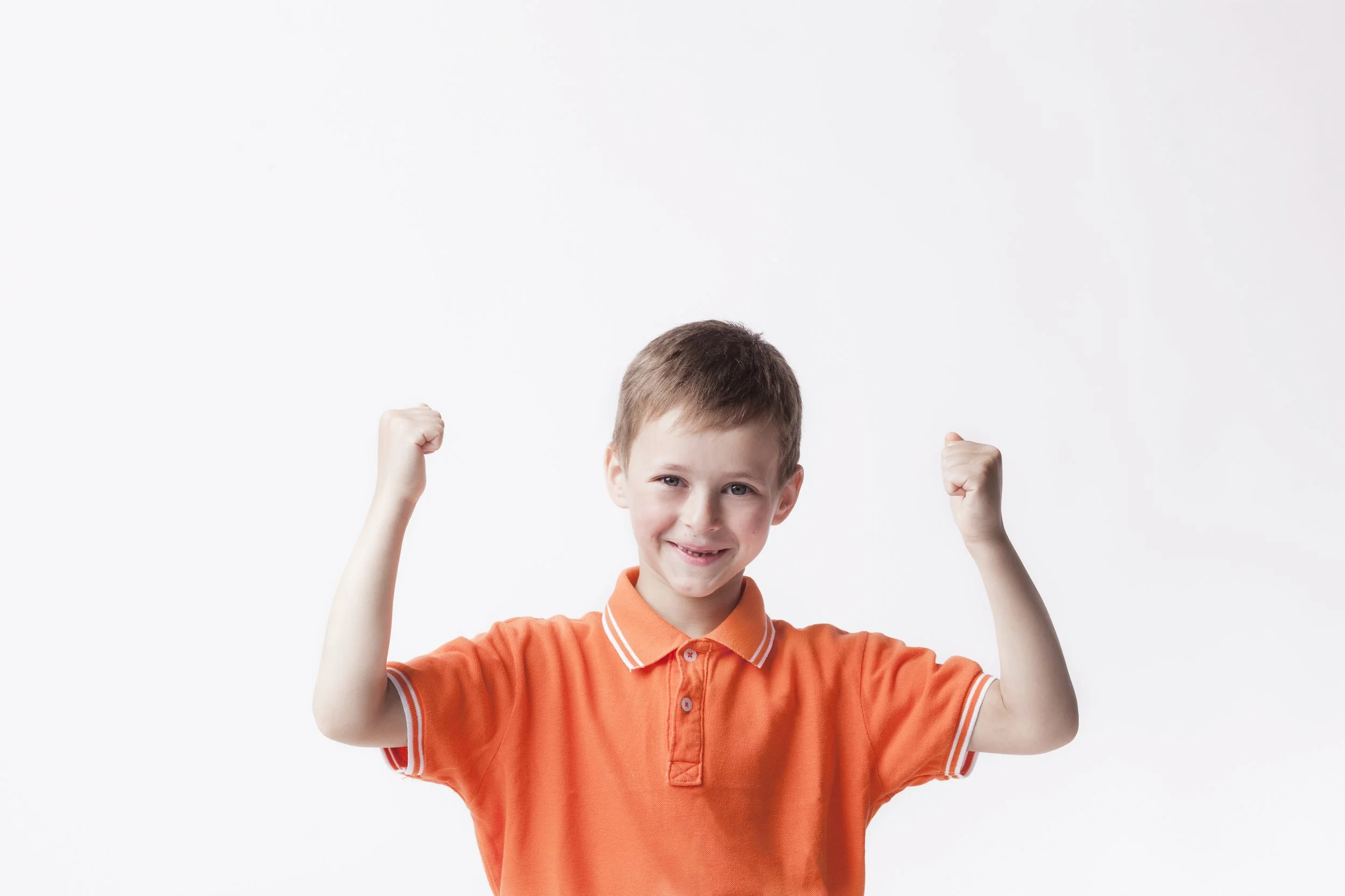 A young boy in an orange polo shirt flexing his biceps with a smile against a plain white background.