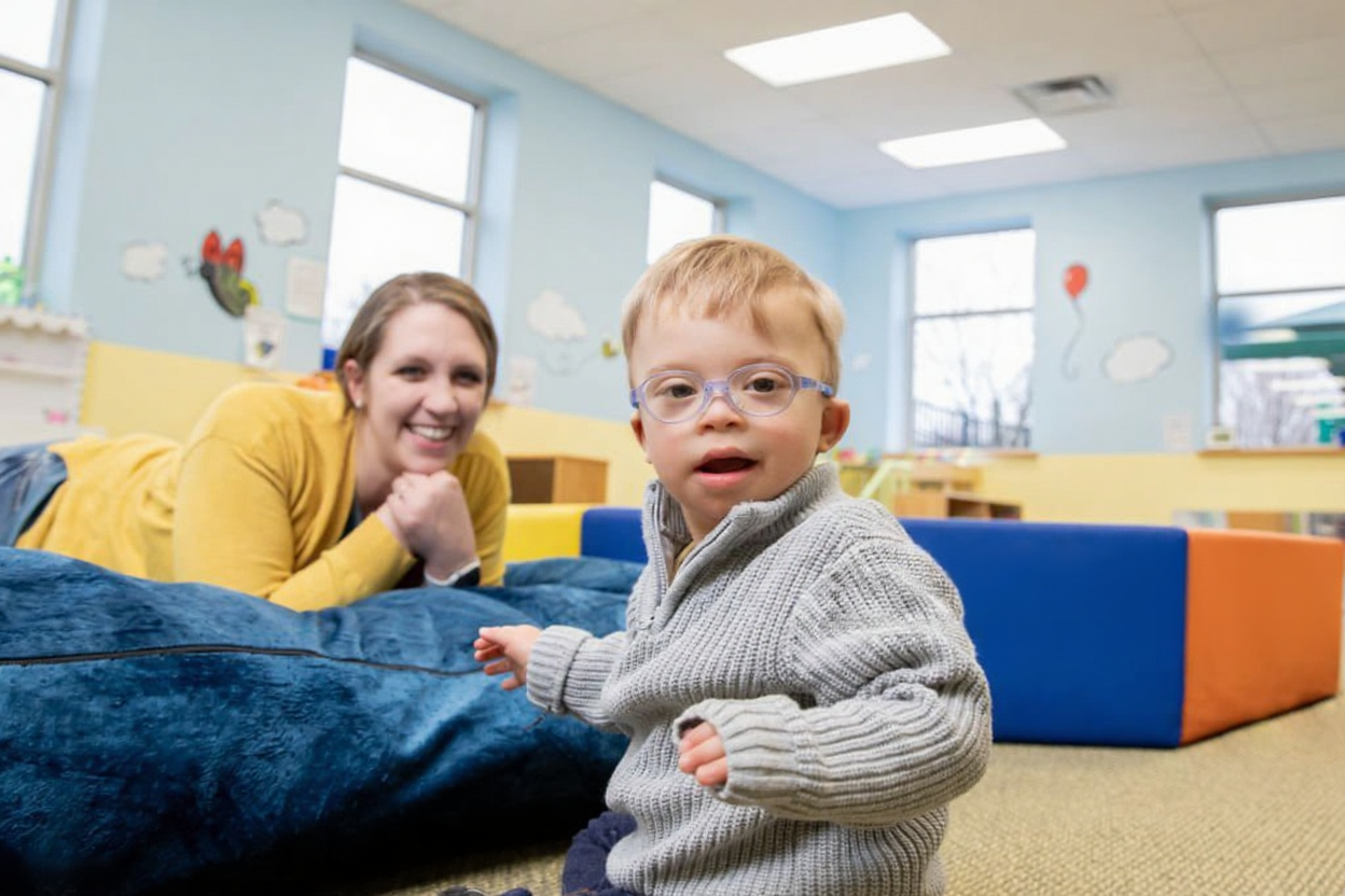 A young boy with glasses and a gray sweater standing in a colorful playroom with a woman lying on a large blue cushion behind him, smiling.