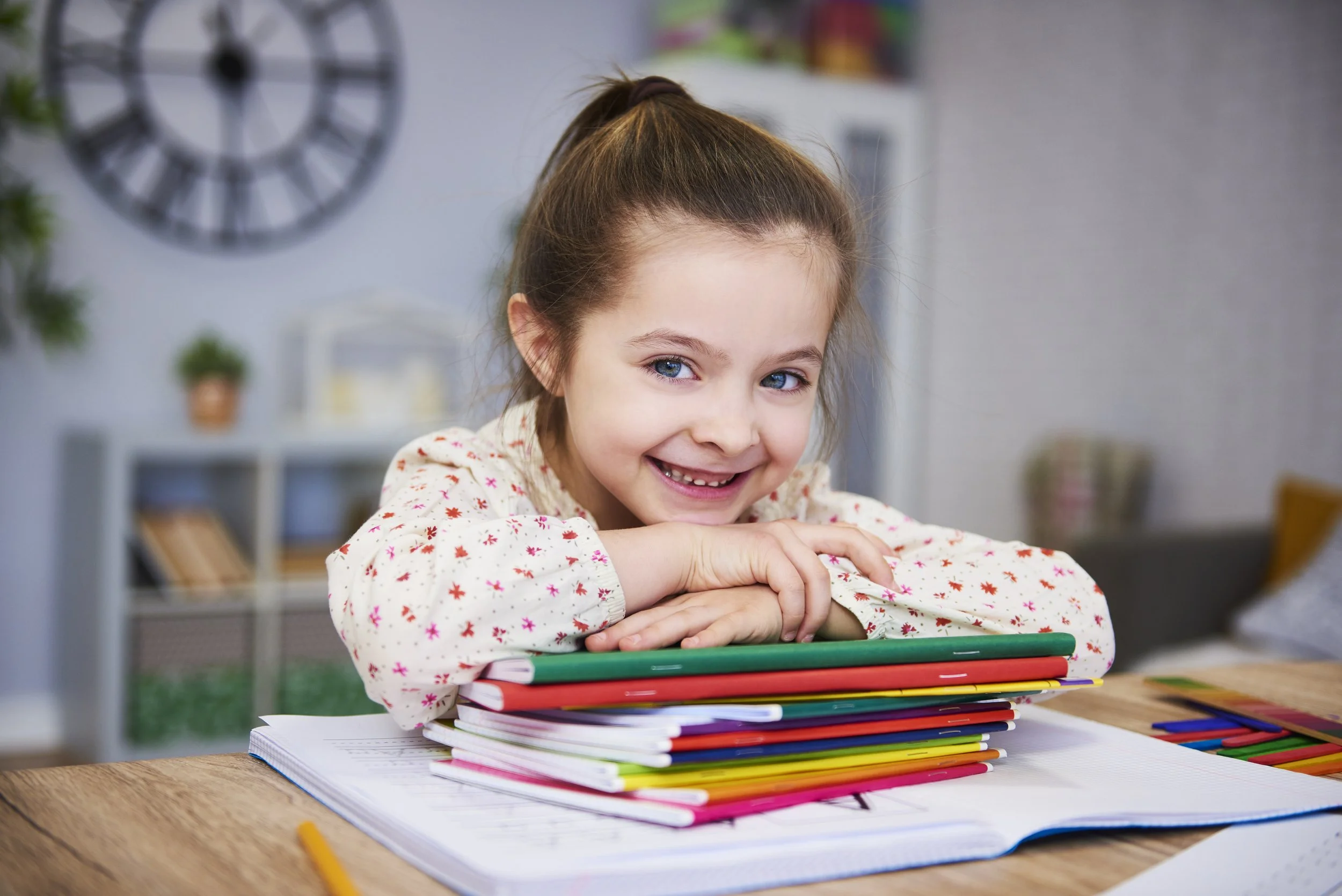 A smiling young girl with blue eyes and brown hair in a ponytail leaning on a pile of color notebooks and coloring supplies on a wooden table.