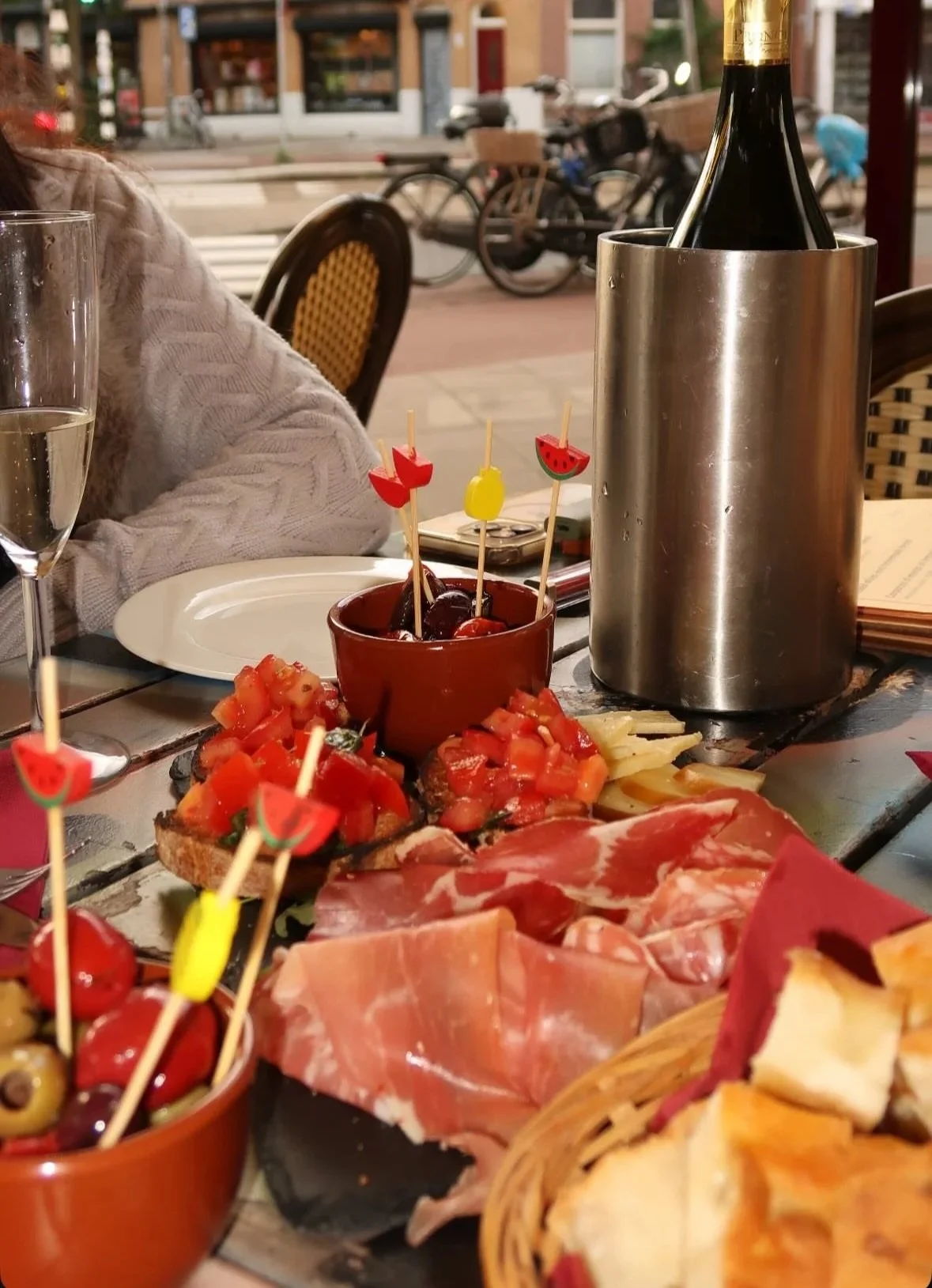 Close-up of a table featuring a variety of appetizers, including cured meats, bruschetta with diced tomatoes, and a bowl of marinated olives with cocktail picks, with a bottle of wine in an ice bucket, and glasses on a patio.
