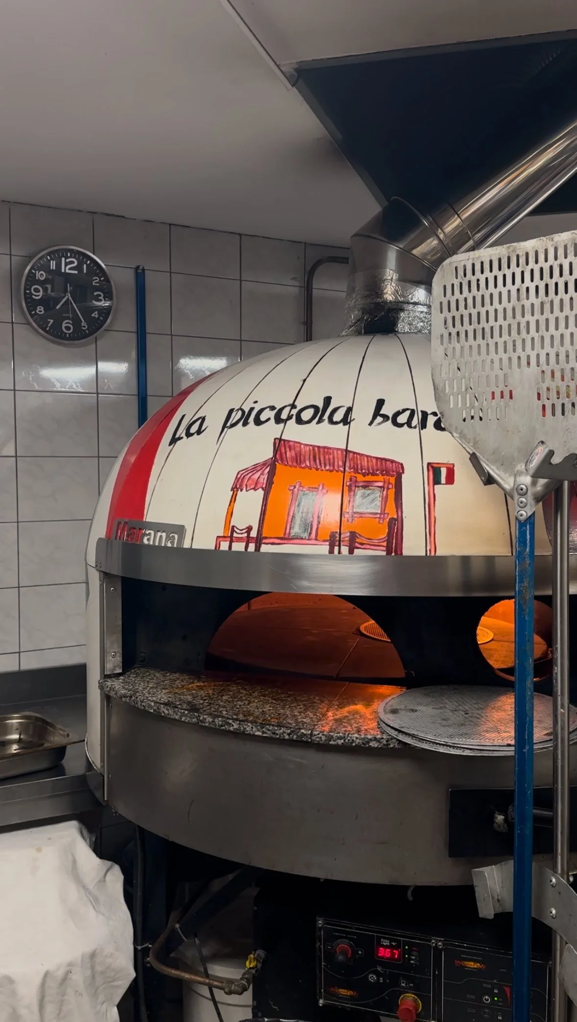 A pizza oven with a dome-shaped top painted with the words 'La Piccola Bar' and an illustration of a small house. The oven is in a commercial kitchen with tiled walls and a clock showing 11:10.