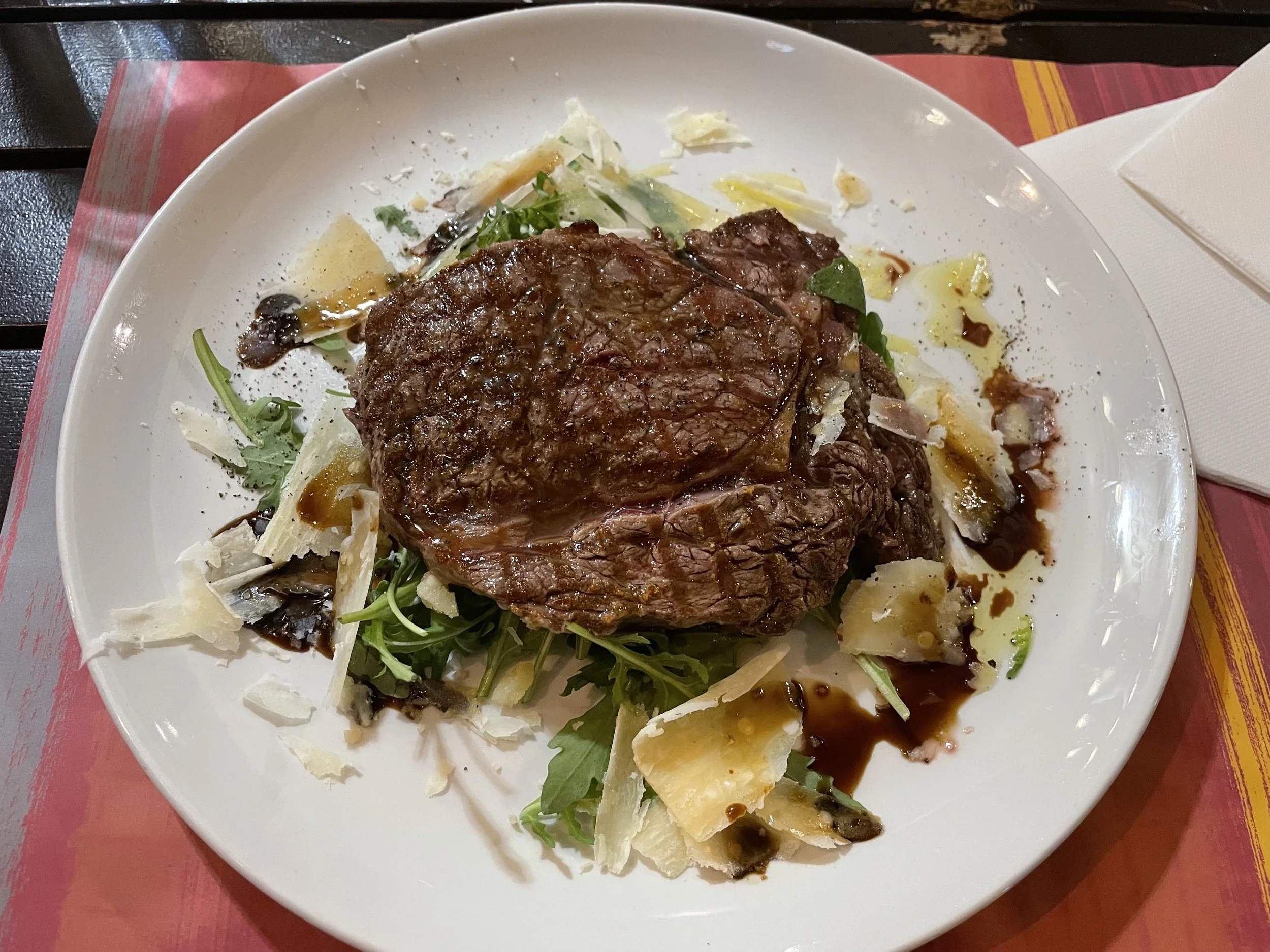 A plate of steak served on a bed of arugula and greens with shaved cheese, drizzled with balsamic glaze and oil.