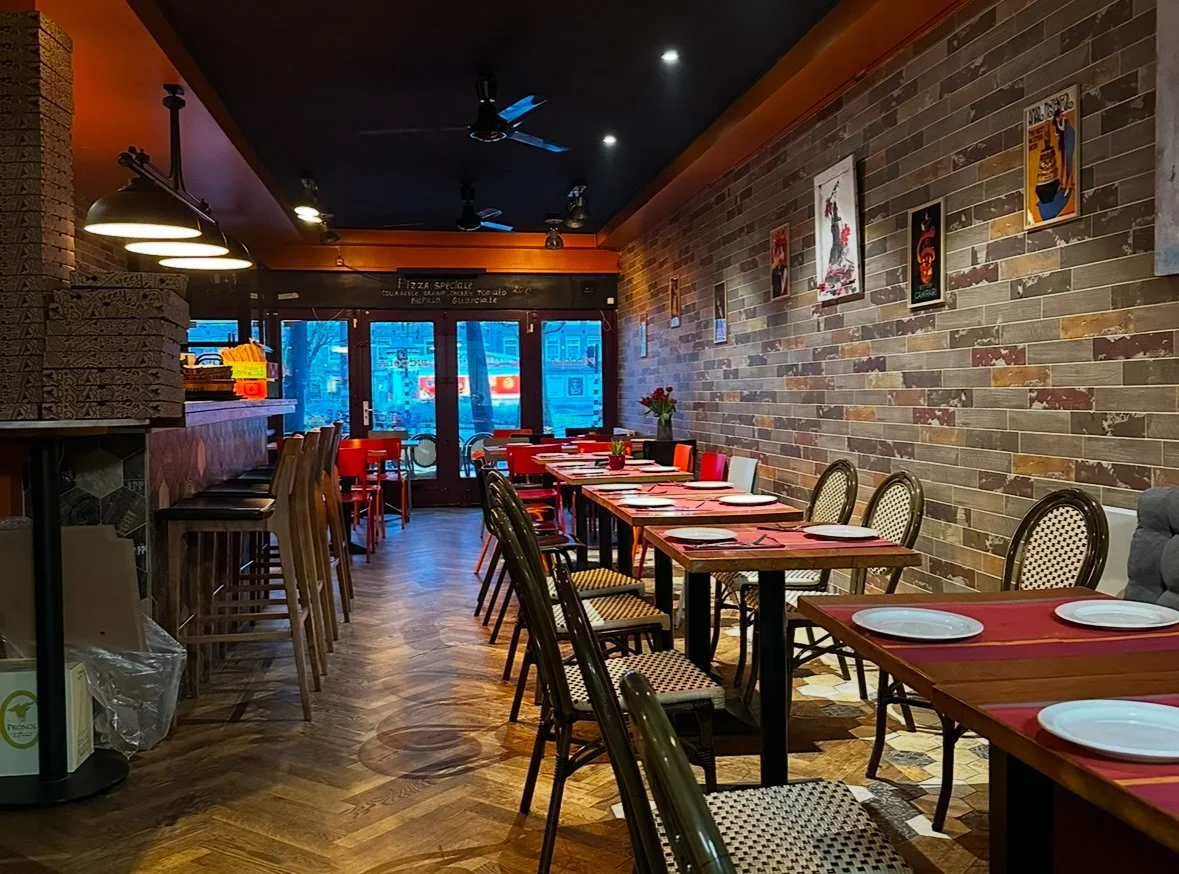 Restaurant interior with rustic brick walls, wooden floors, and tables set with plates and napkins, decorated with framed artwork on the wall.
