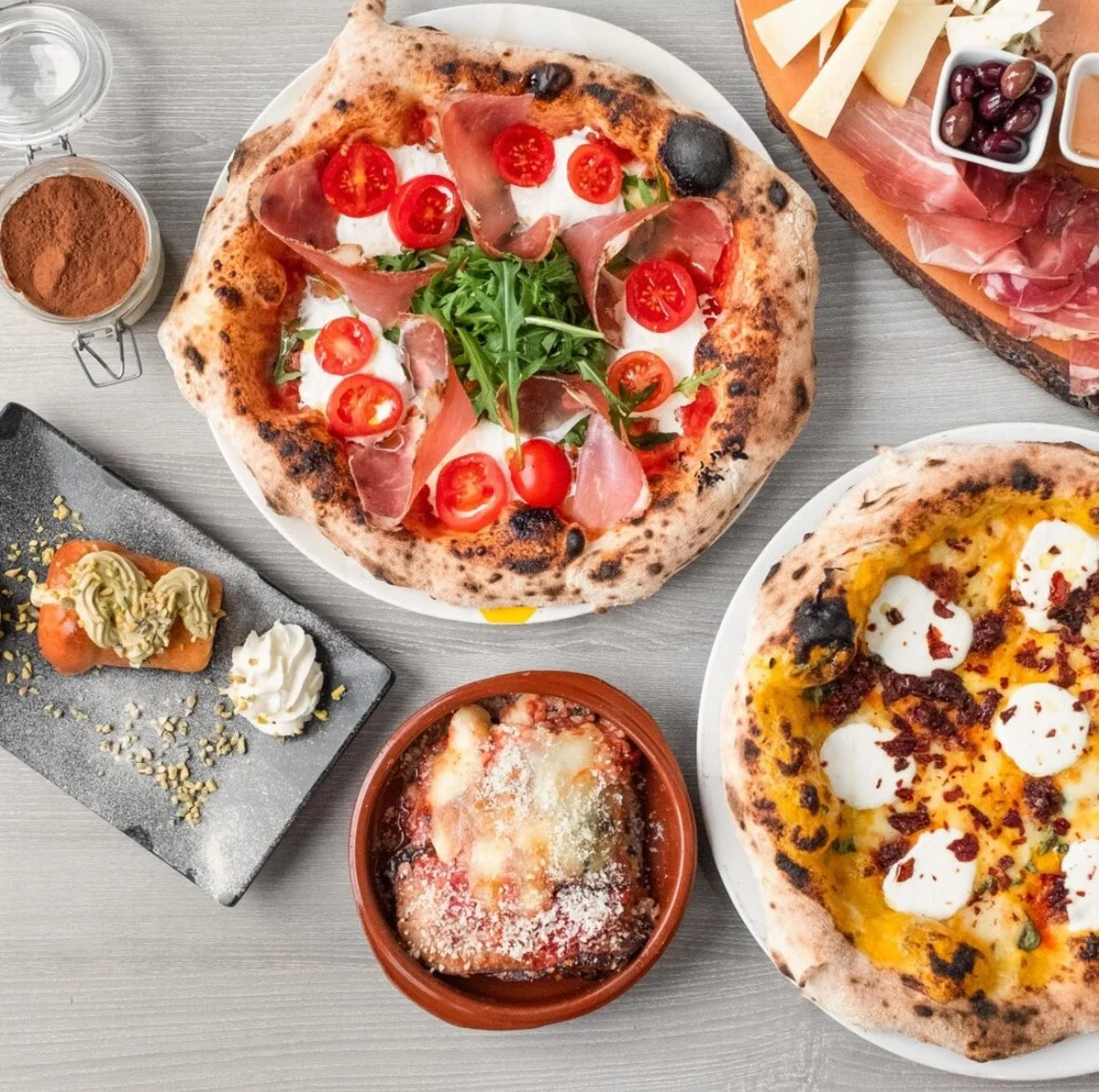 Assorted pizzas, a small dish of olive tapenade, a plate with spring rolls, and small bowls of olives and sauces on a gray table.