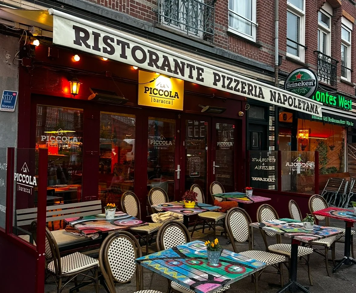 Outdoor seating area of an Italian restaurant called La Piccola in Napoli, with colorful tables, potted plants, and building signage with banners for risorante, pizzeria, and Heineken.