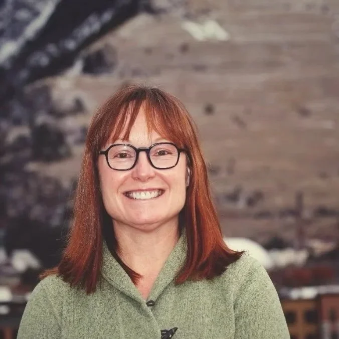 A woman with short gray hair wearing glasses, a dark blazer, and a floral shirt, smiling against a dark background.