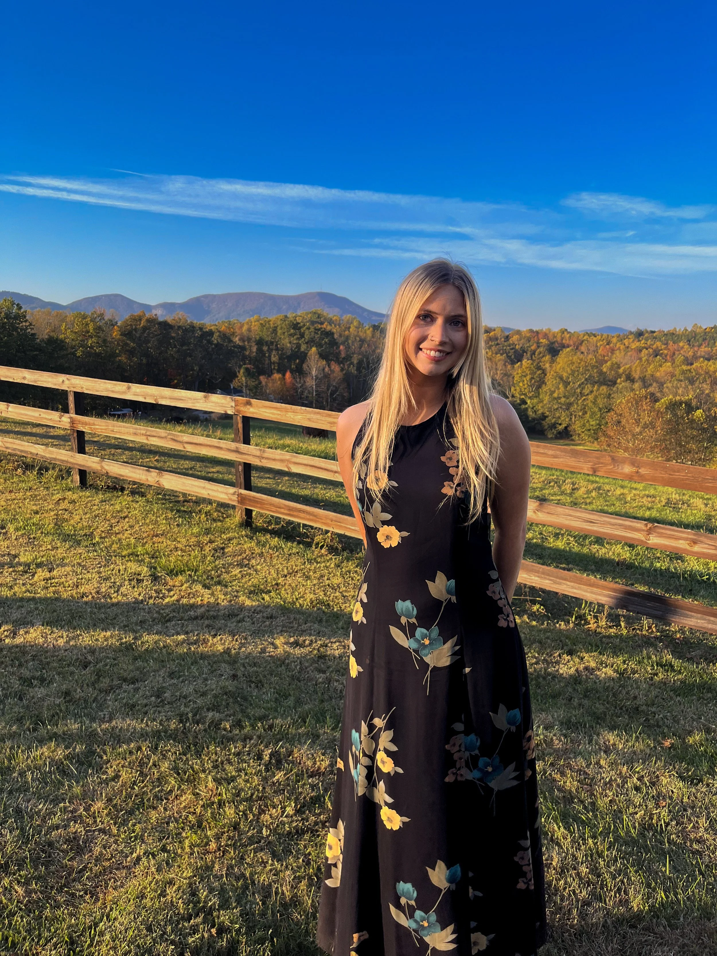 A young woman with long blonde hair smiling outdoors in a black floral dress with yellow and blue flowers, standing next to a wooden fence with a scenic mountain view in the background during sunset.