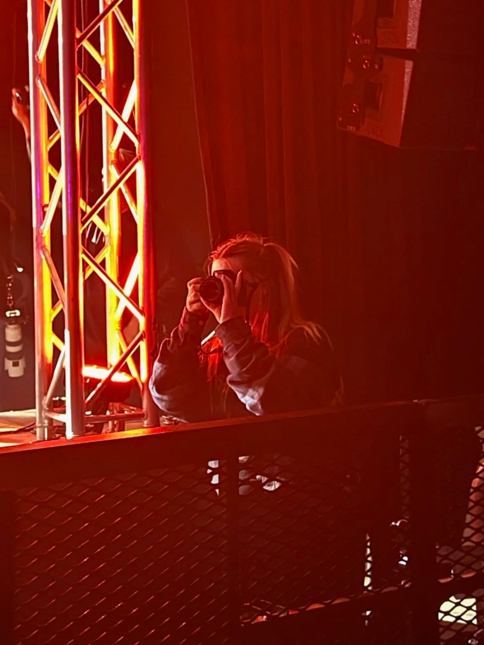 Person taking a photo in front of a colorful stage setup with metal trusses and red lighting.