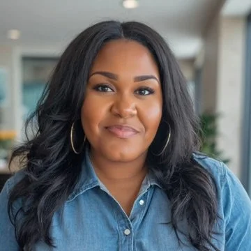 A woman with long black hair, wearing hoop earrings and a denim jacket, smiling softly in an indoor setting.