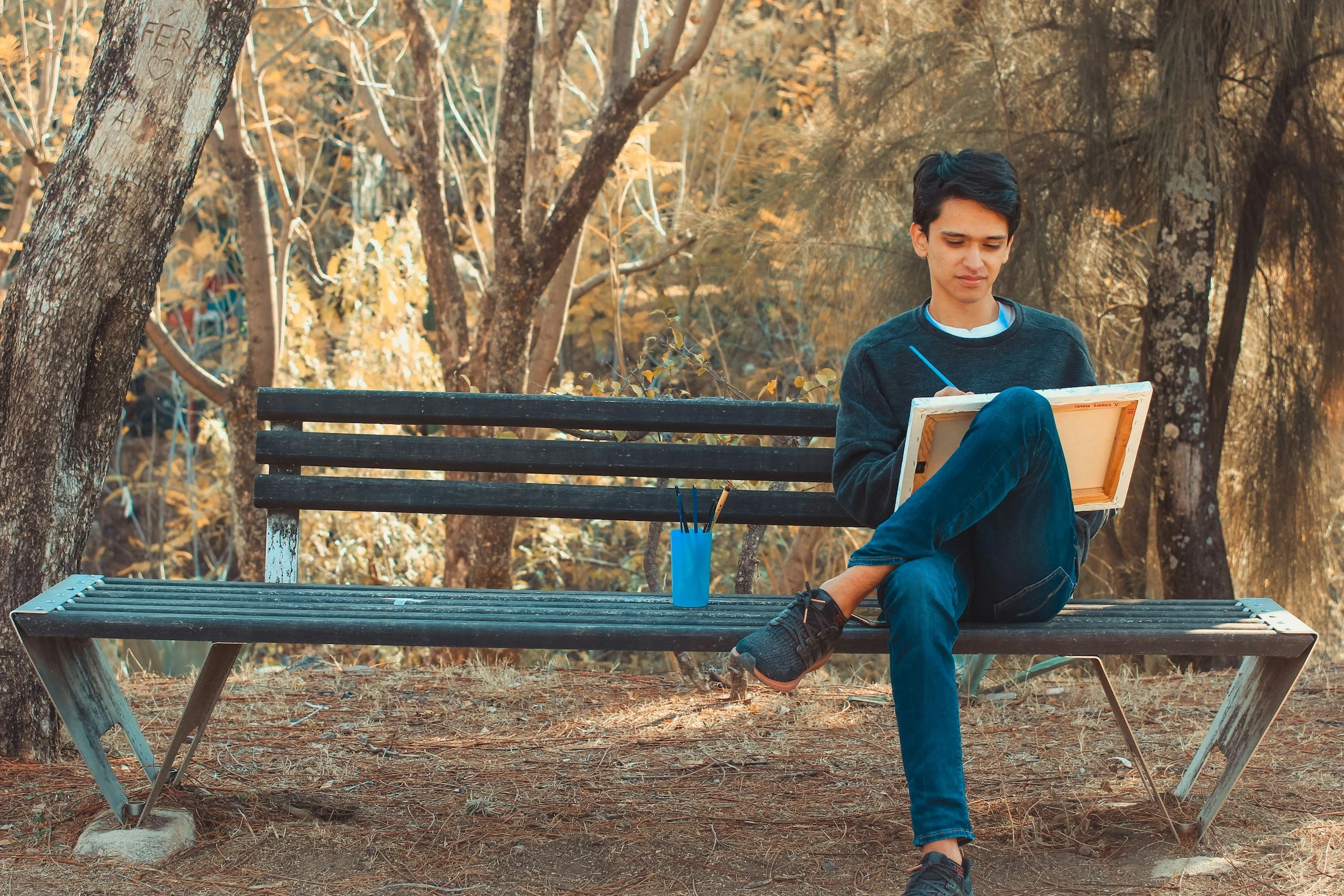 A young man sitting on a park bench outdoors, holding a sketchpad and drawing with a pencil. There are trees in the background with autumn-colored leaves.