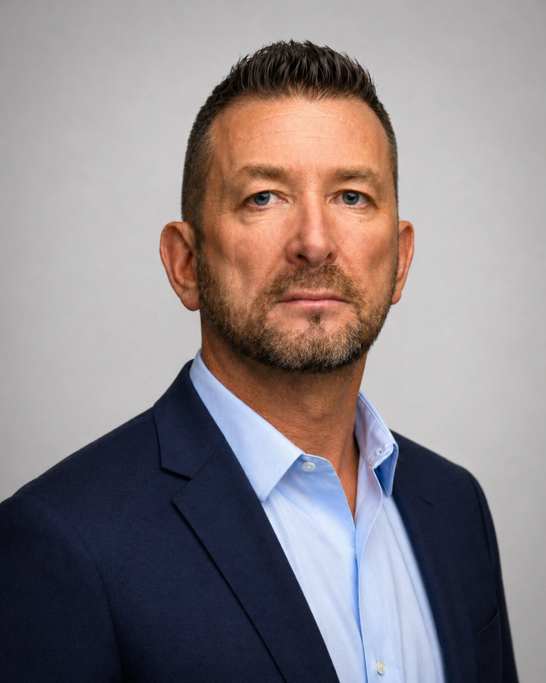 A professional headshot of a middle-aged man with short brown hair, a beard, wearing a dark navy suit and a light blue collared shirt, against a plain gray background.