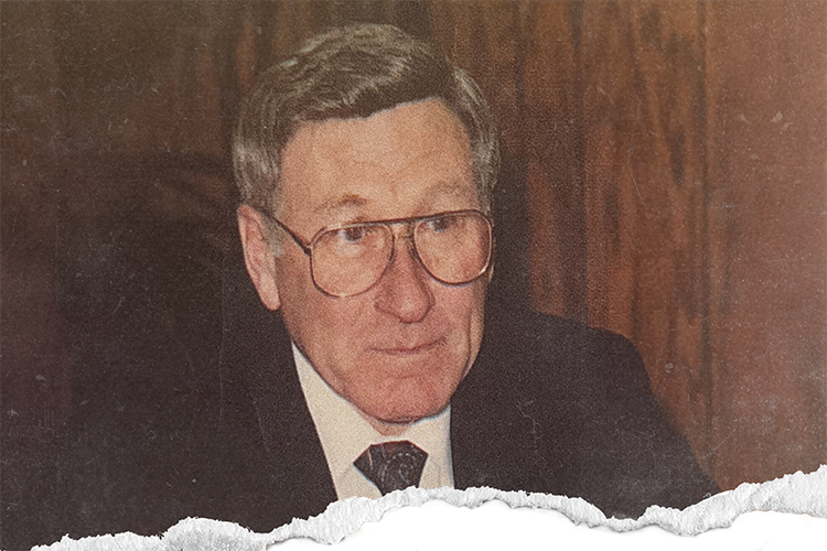 An older man with glasses wearing a suit with wood panel wall behind him