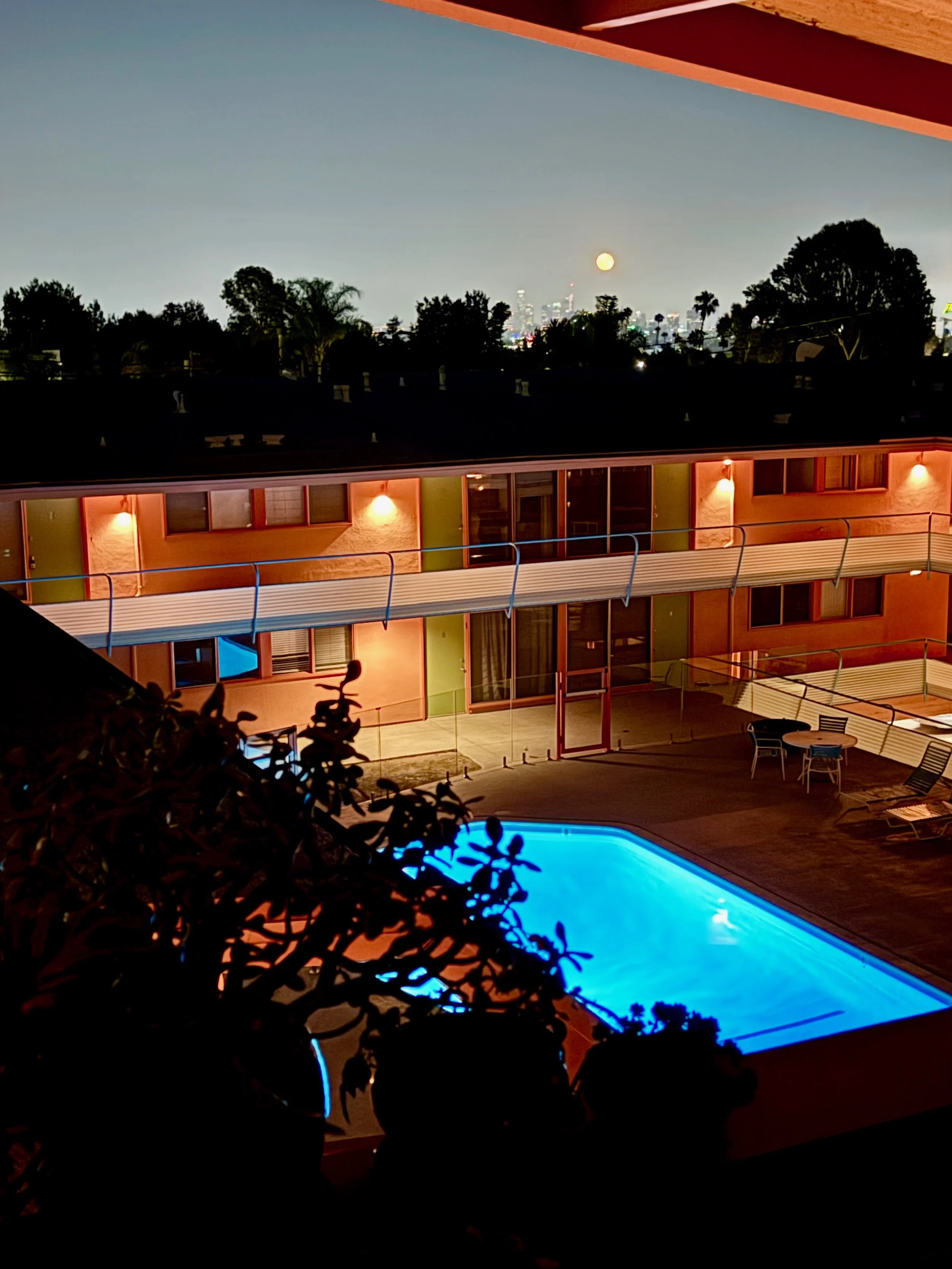 Night view of a lit swimming pool with a building in the background, city skyline, and full moon in the sky.