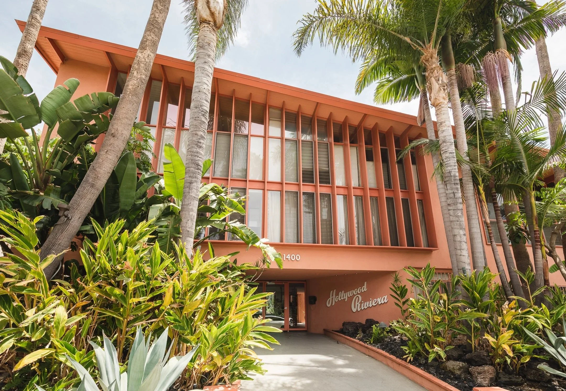 Pink-colored modern building with tall, narrow windows and the sign 'Hollywood Riviera' at the entrance, surrounded by lush tropical plants and tall palm trees.