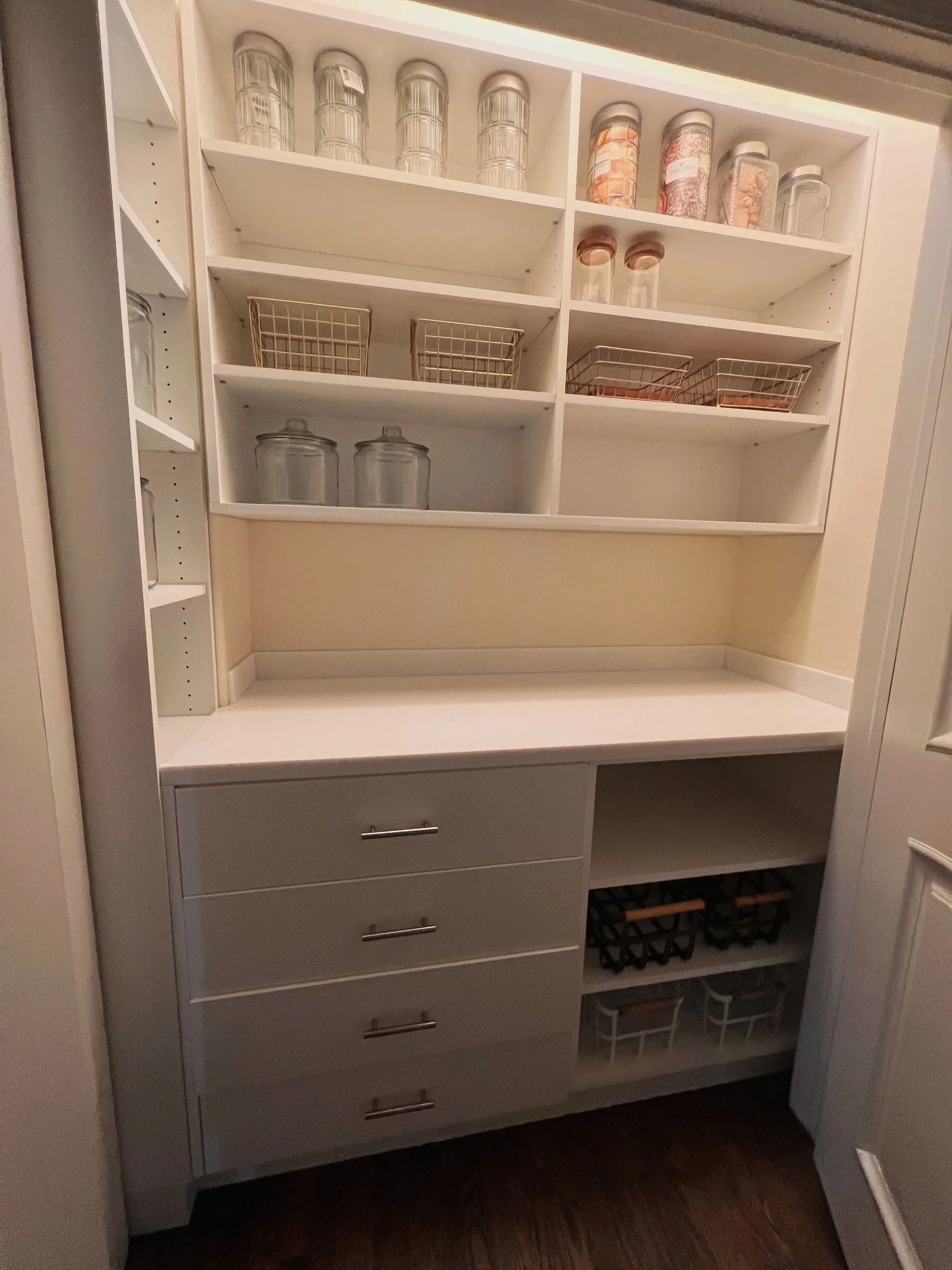Organized white pantry with jars and baskets on open shelving above drawers
