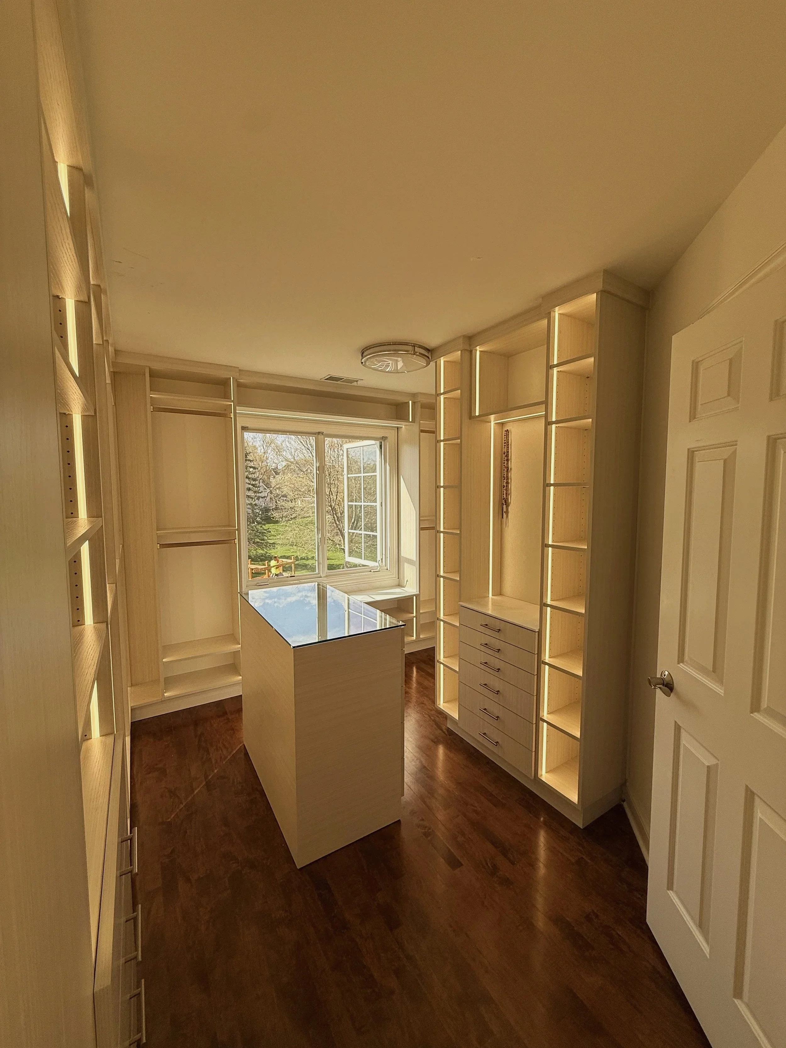Warm-tone custom walk-in closet with center island, window view, and vertical LED lighting