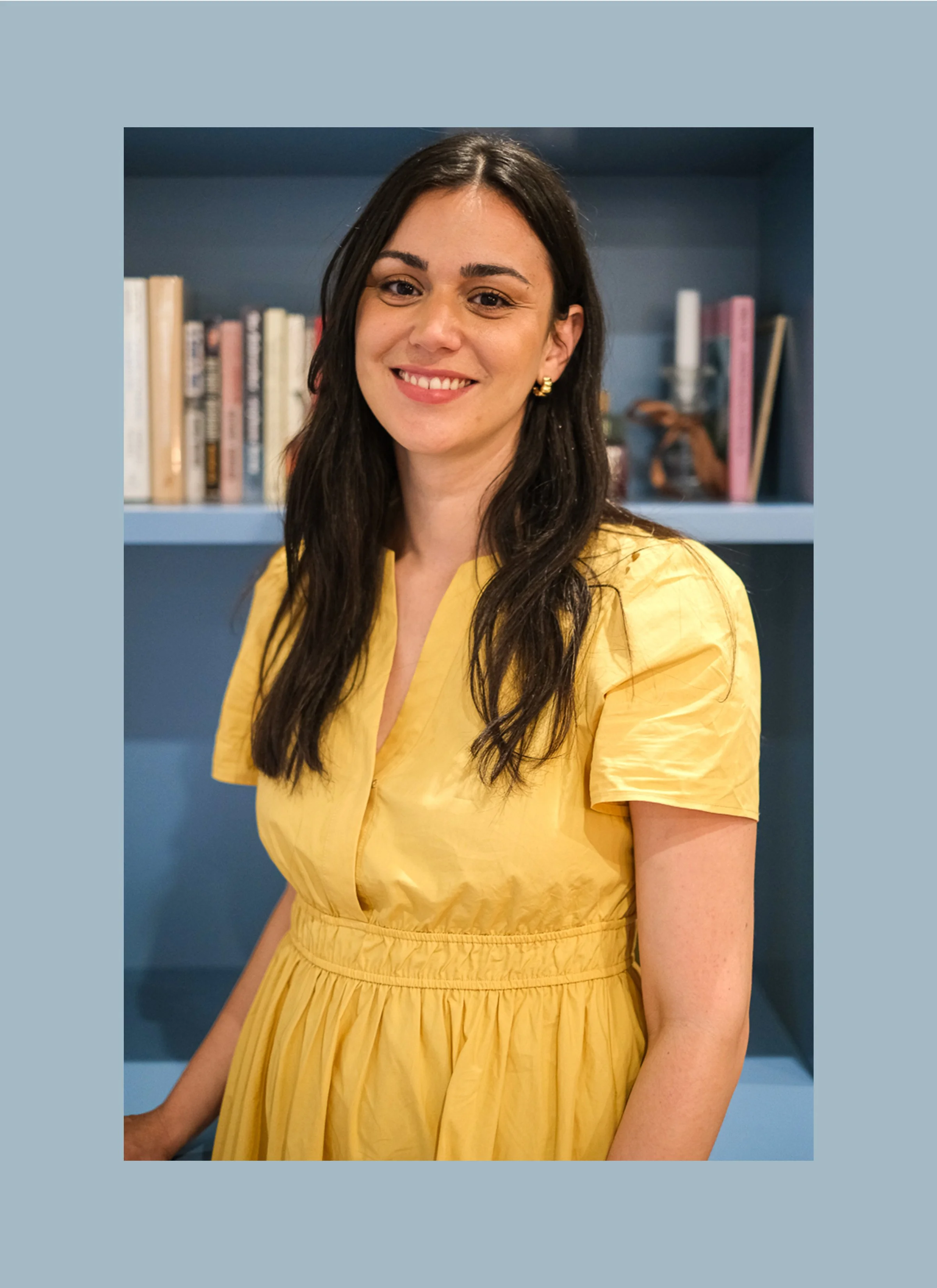 A woman with long dark hair, wearing a yellow dress, smiling and standing in front of a bookshelf with books and decorative objects.