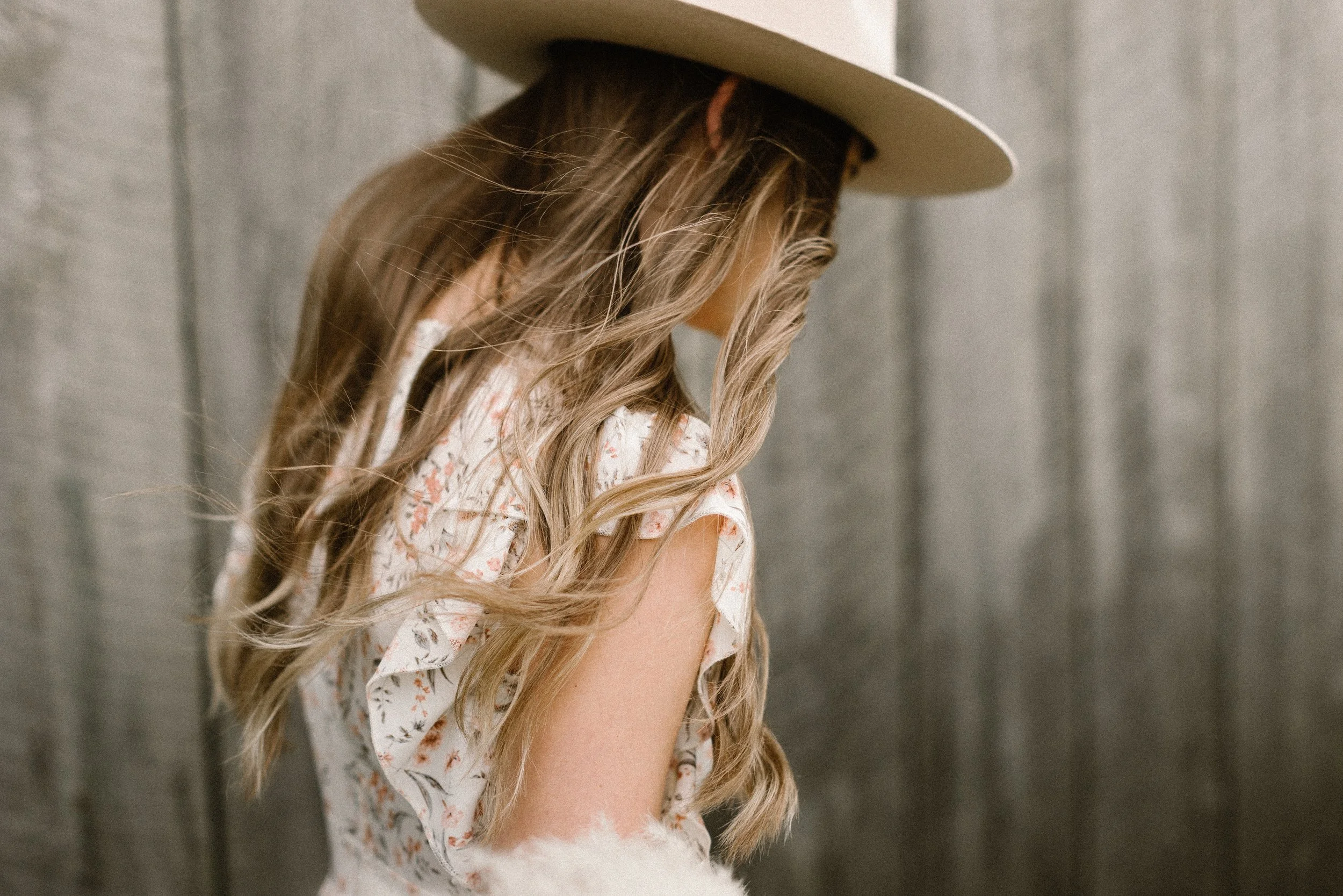 A woman with long, wavy blonde hair wearing a beige wide-brimmed hat and a floral dress, standing against a gray wooden background.