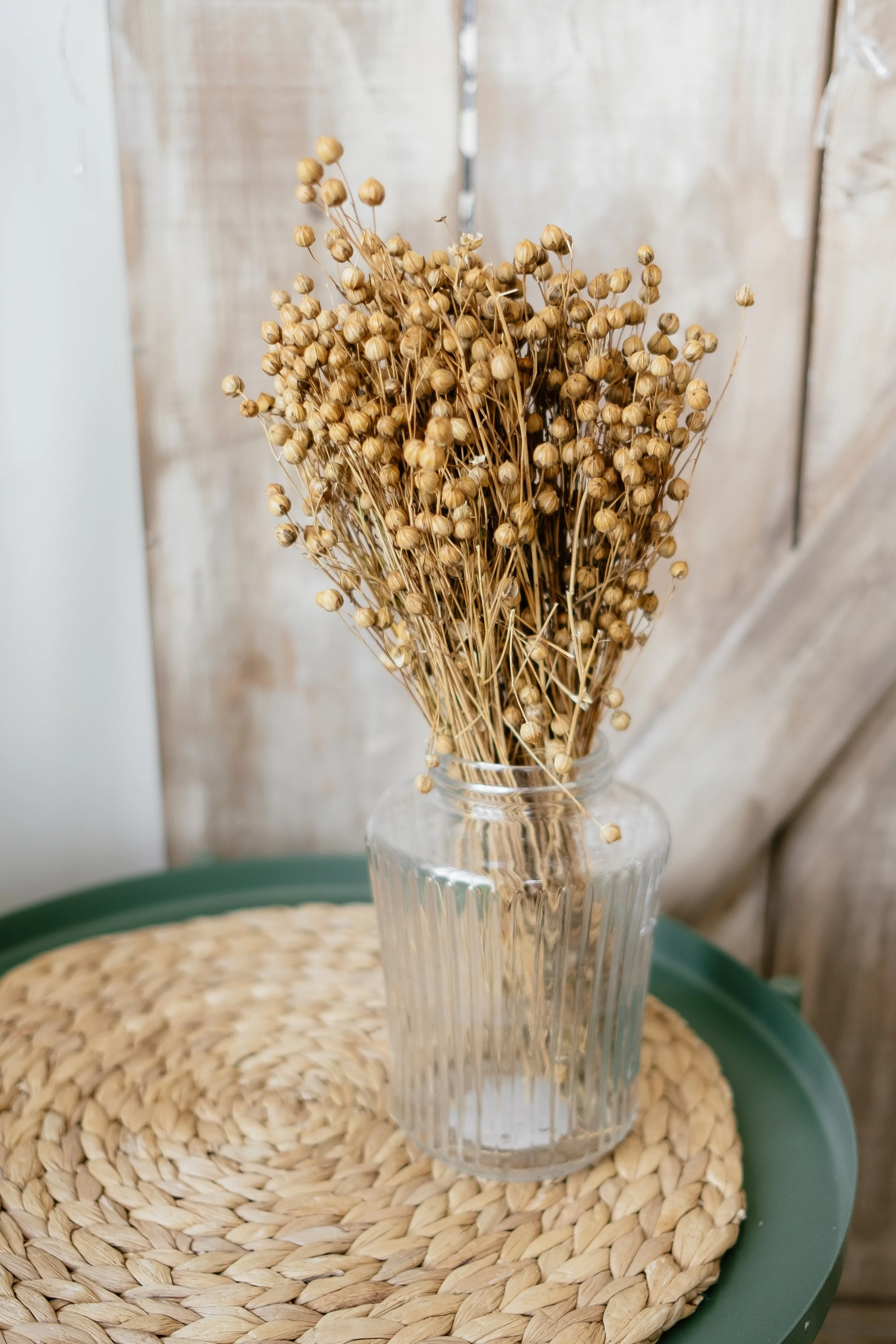 A clear glass vase filled with dried beige seed pods and stems, placed on a woven circular placemat on a green table against a wooden wall background.
