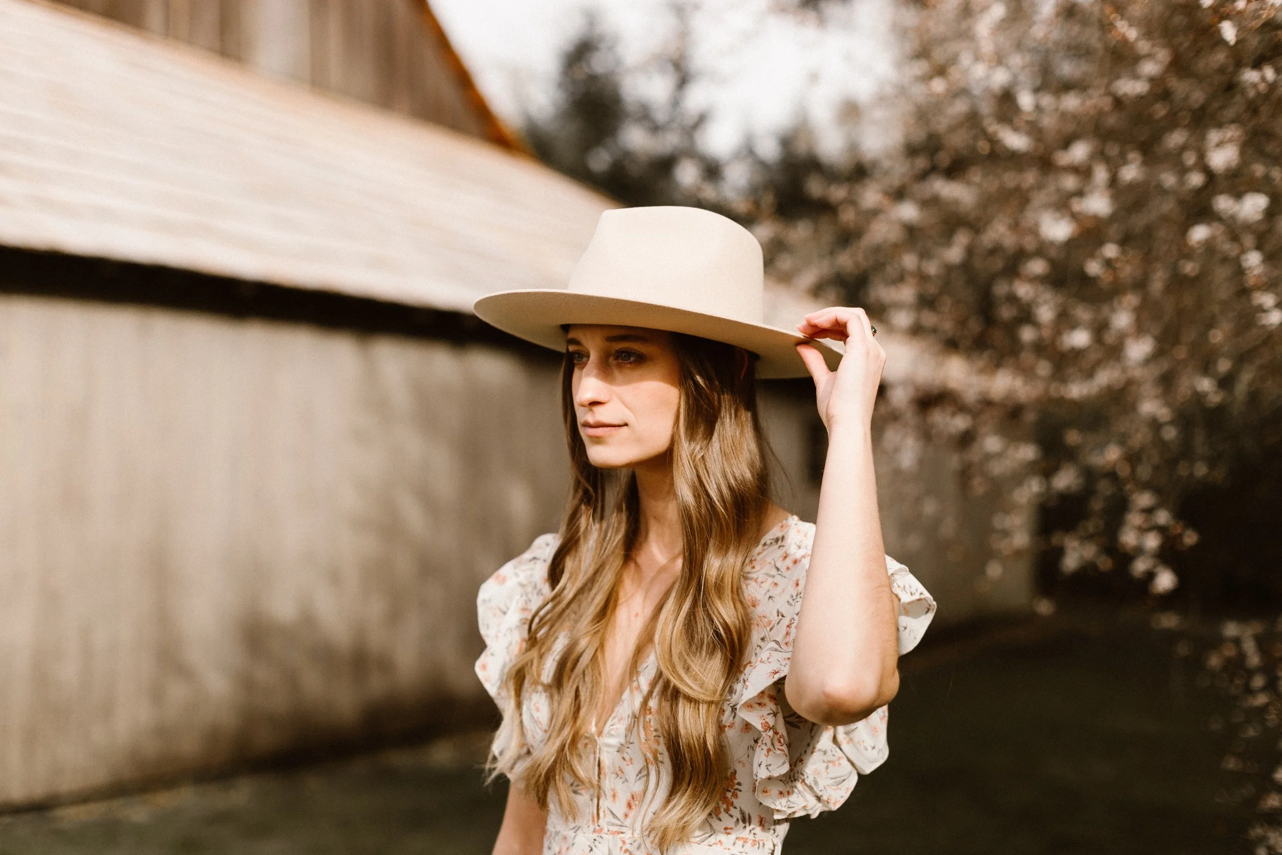 A young woman with long wavy hair wearing a floral dress and a wide-brimmed hat, standing outdoors near a fence and blooming trees.