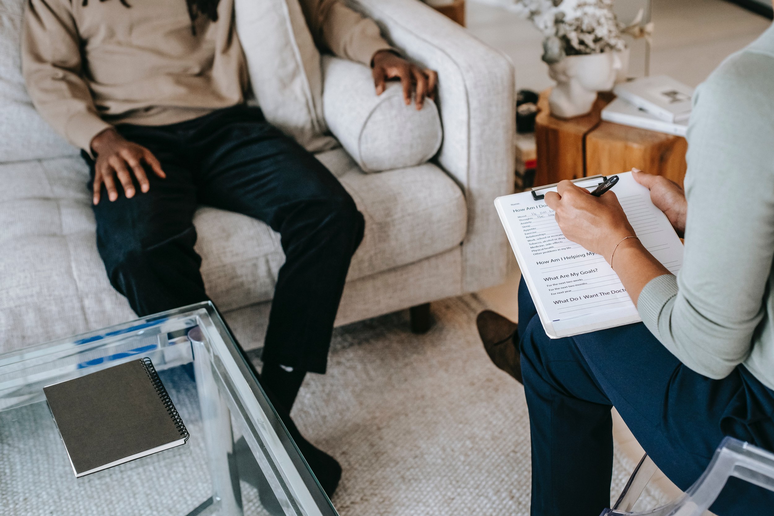 A person sitting on a beige sofa during a medical consultation with a therapist or doctor who is taking notes on a clipboard.