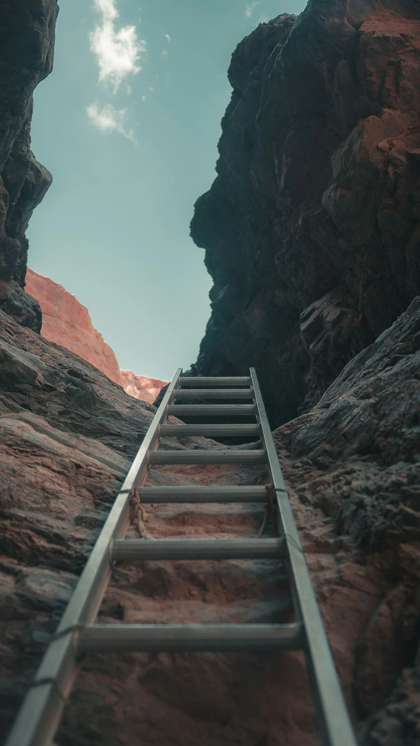 A metal ladder leaning against a rocky canyon with a view of the sky and clouds above.