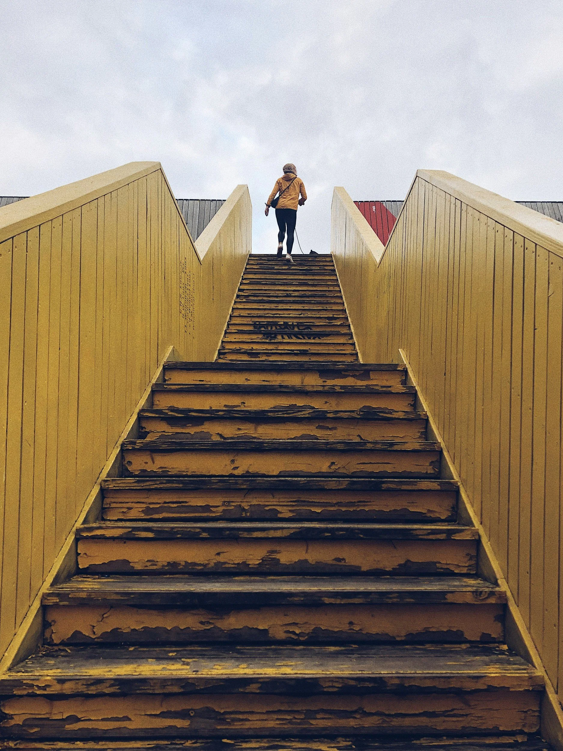 A person walking down a weathered wooden staircase with yellow railings, leading upward against a cloudy sky.