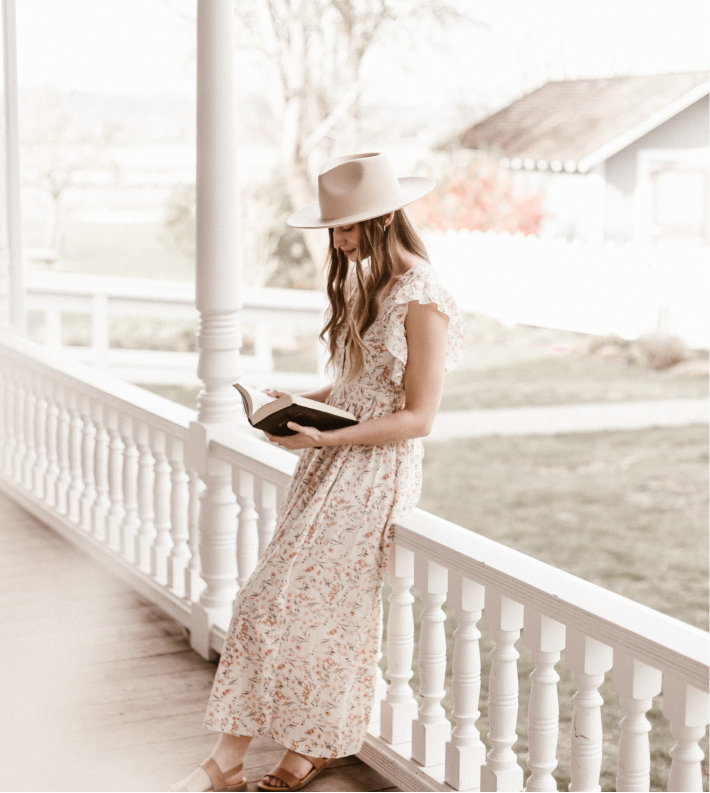A woman wearing a wide-brimmed hat and a long floral dress stands on a porch, leaning against the white railing while reading a book.