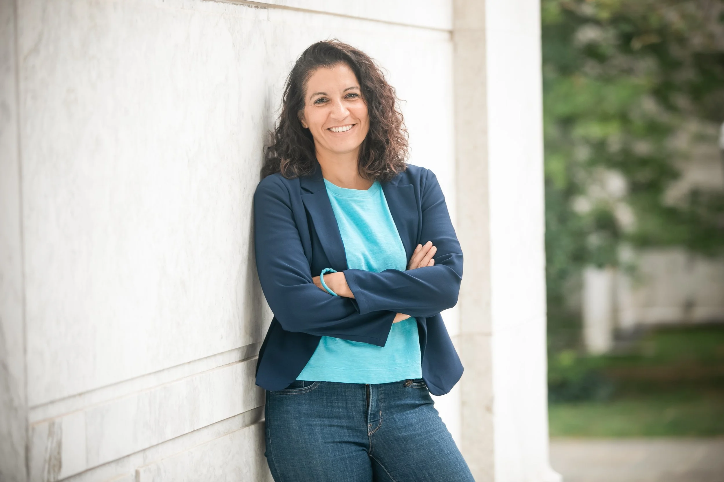 A woman smiling and standing with arms crossed against a light-colored wall outdoors, wearing a blue blazer and jeans.