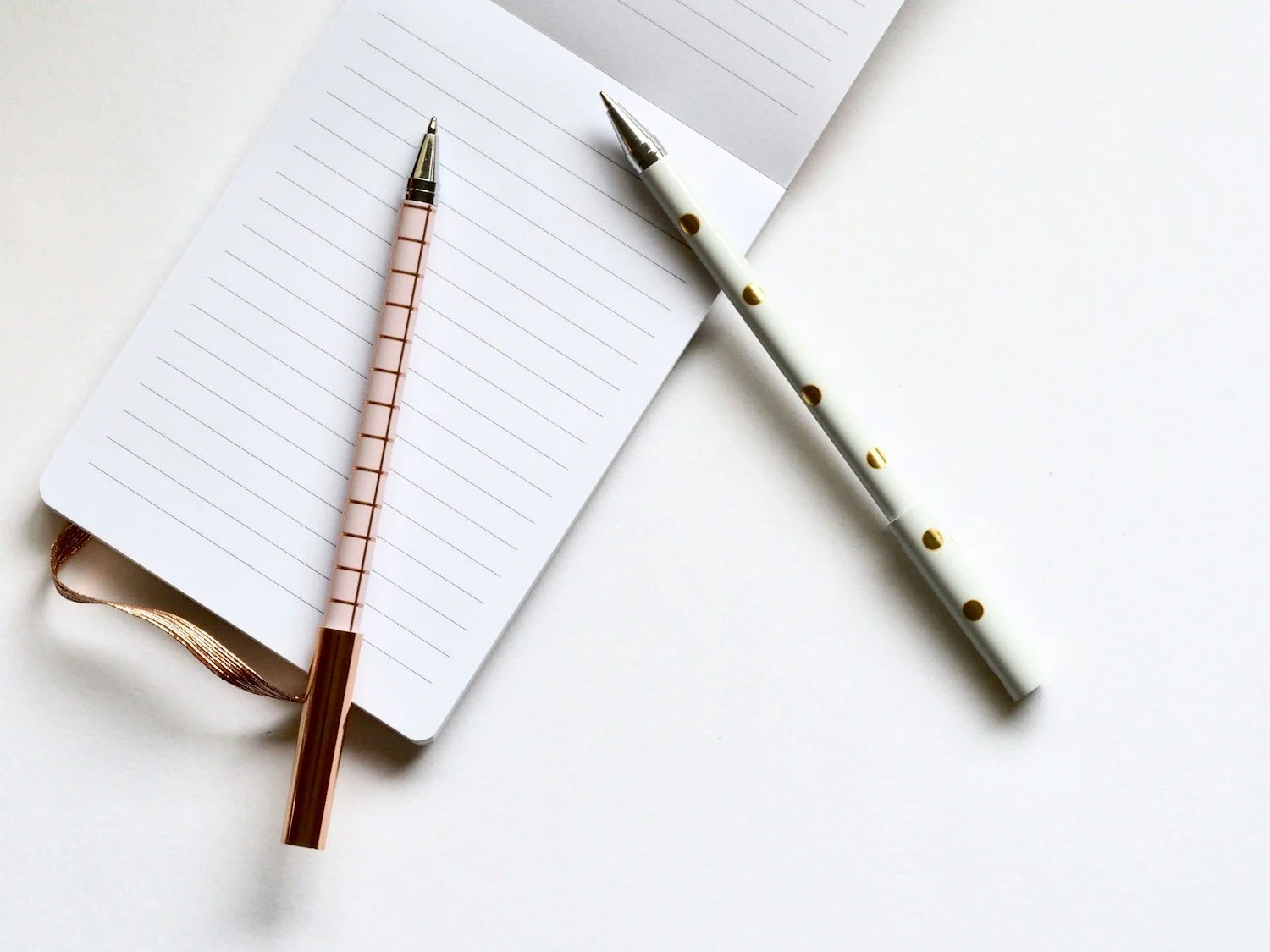 A pink and gold pen resting on an open, lined notebook with a white and gold pen nearby on a white surface.