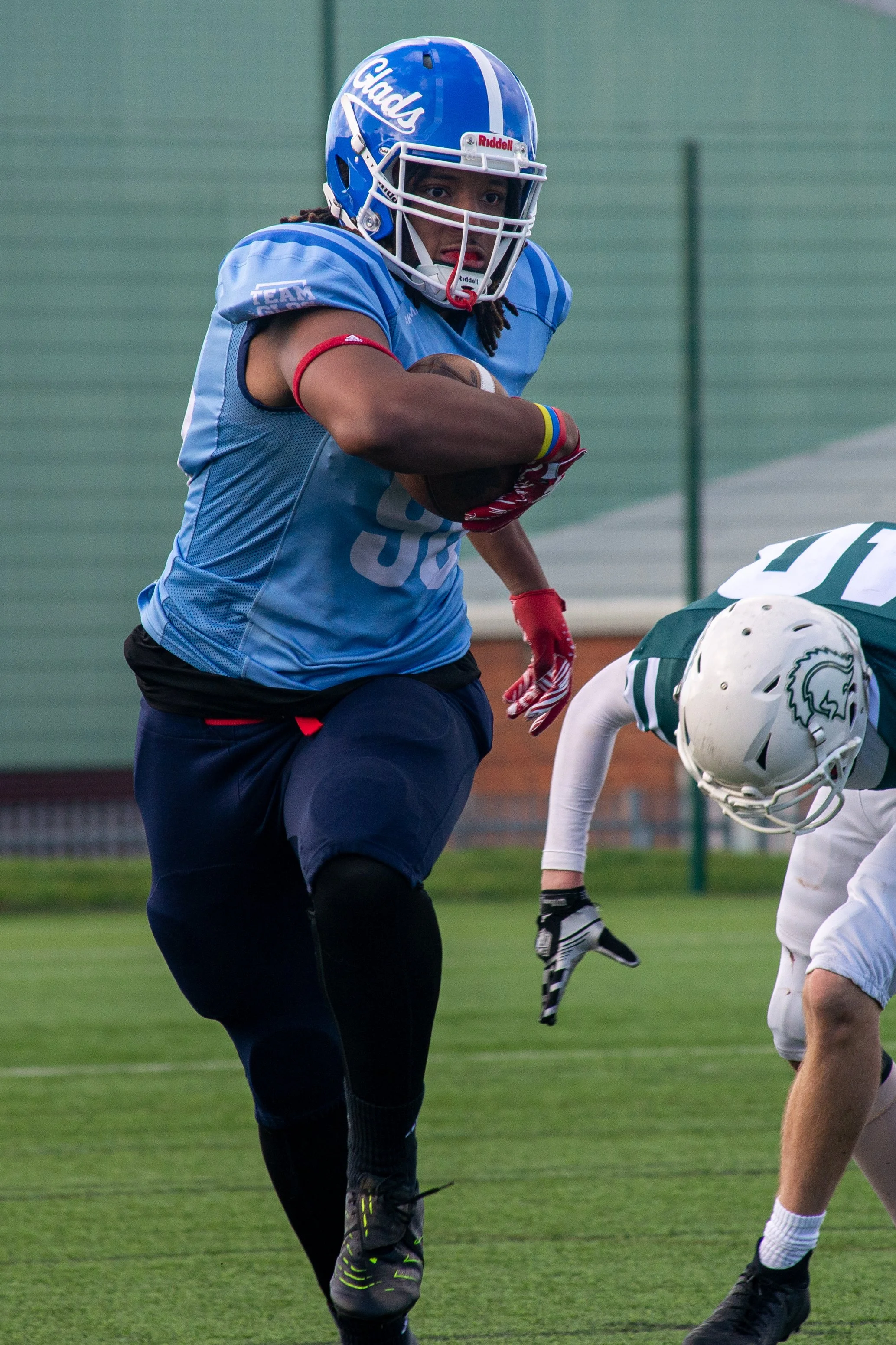 A football player in a blue jersey and helmet catching a football during a practice or game, with opponents in white and green jerseys nearby.