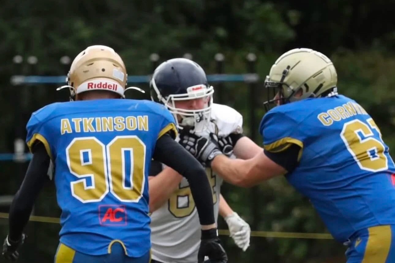 Two football players in blue jerseys with yellow numbers and gold helmets are blocking a player in a white jersey and black helmet during a game.