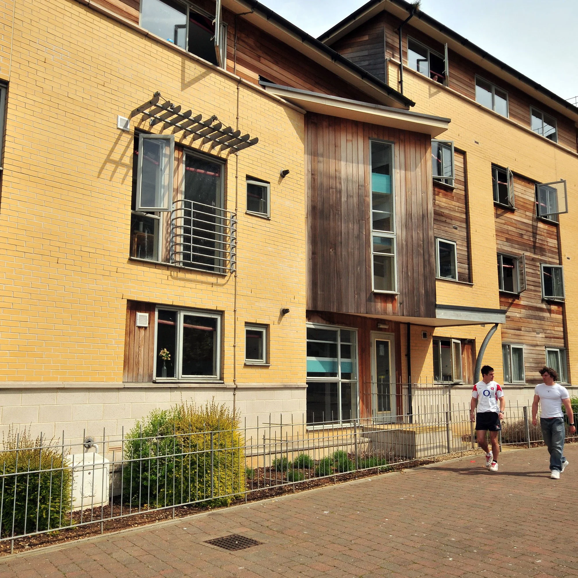 Modern multi-story apartment building with yellow brick and wood paneling, several open windows, and two young men walking on a paved sidewalk in front.