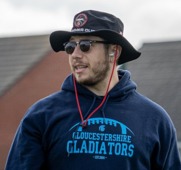A man outdoors wearing sunglasses, a wide-brimmed hat with logos, and a navy hoodie with 'Gloucestershire Gladiators' written on it, with a blurred building in the background.