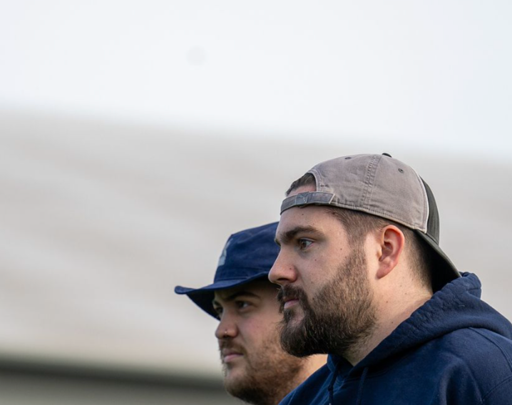 Two men with serious expressions standing outdoors, wearing hoodies and hats, background of a cloudy sky.